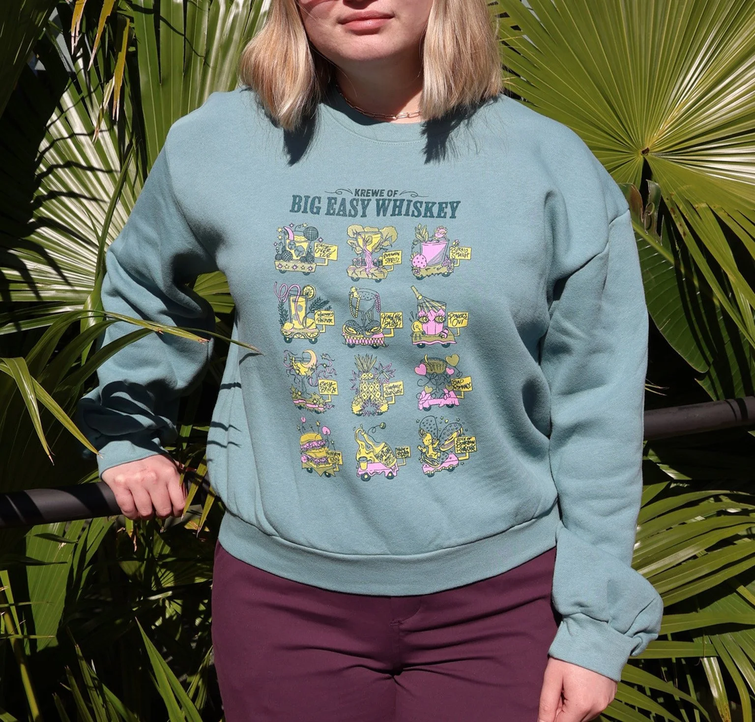 A woman wearing a blue sweatshirt with a colorful graphic and the text 'Krewe of Big Easy Whiskey' stands outdoors in front of large green palm leaves.