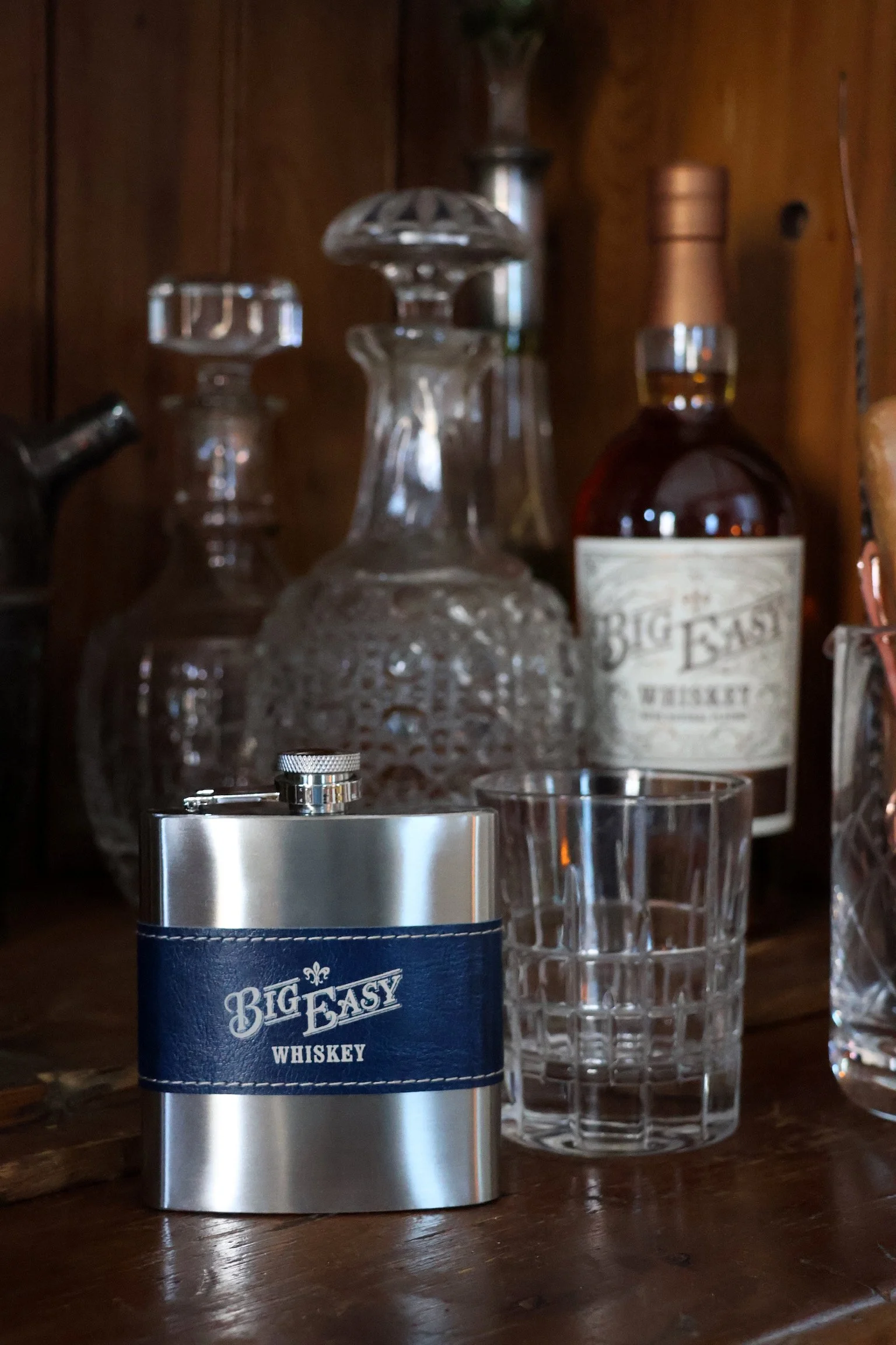 A metal flask labeled 'Big Easy Whiskey' next to a glass on a wooden bar or table, with glass decanters and a bottle of whiskey in the background.