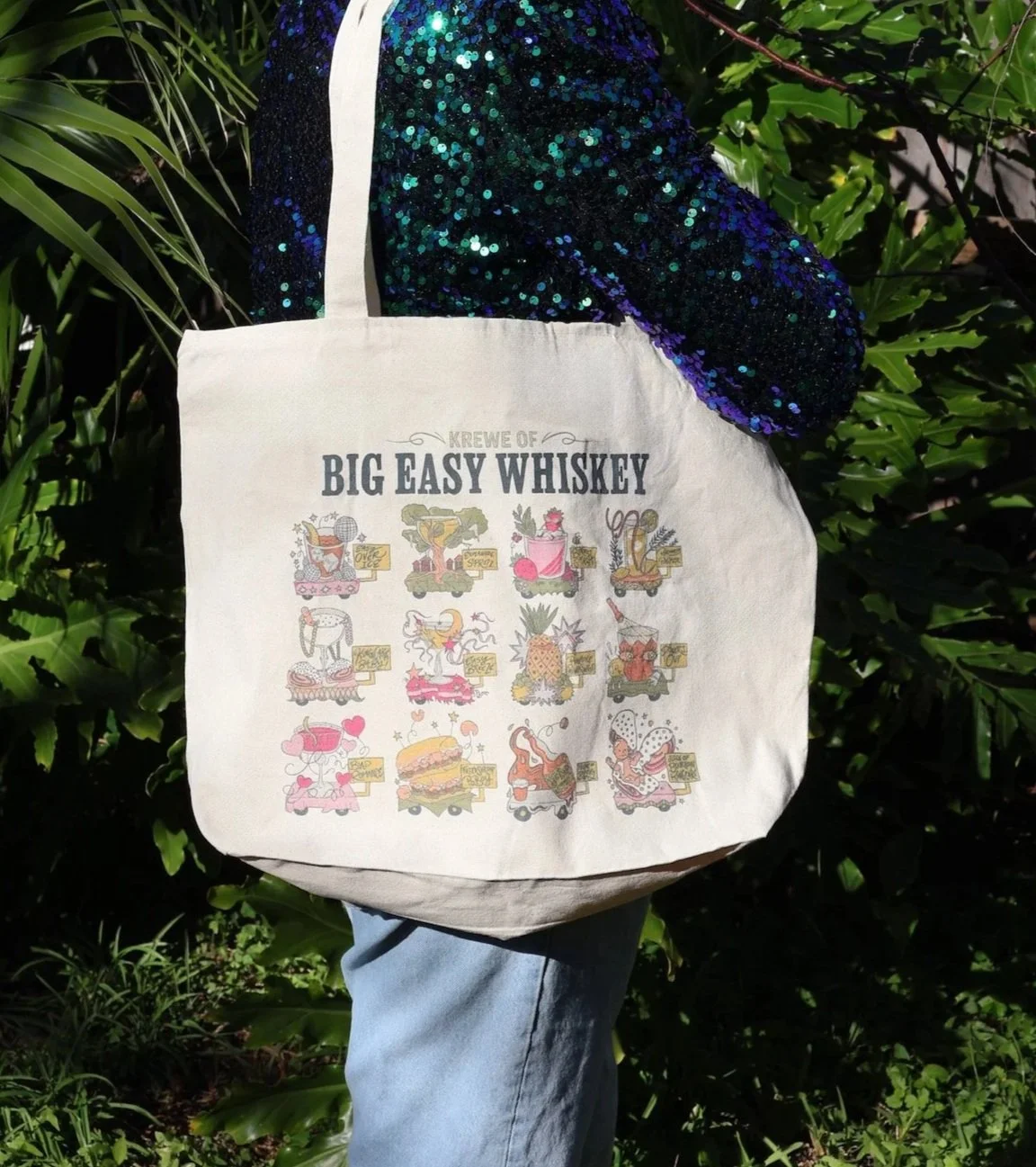 A white tote bag with colorful cartoon graphics and the text "KREWE OF BIG EASY WHISKEY" on it, hanging against green foliage. The person carrying it is wearing a shiny, sequined blue jacket and light blue jeans.