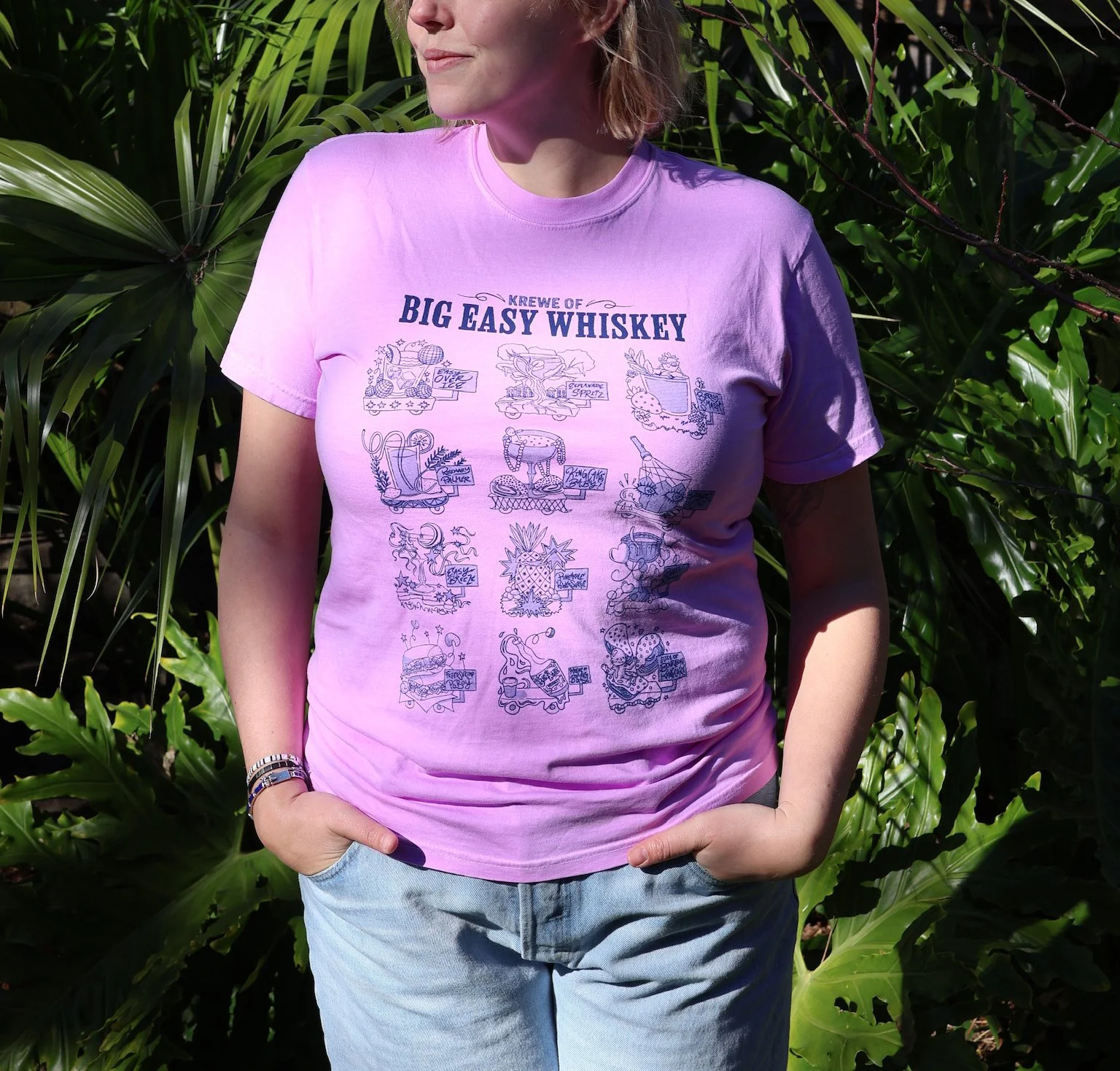 A woman wearing a purple T-shirt with illustrations and text, standing outdoors among green plants with her hands in her pockets.