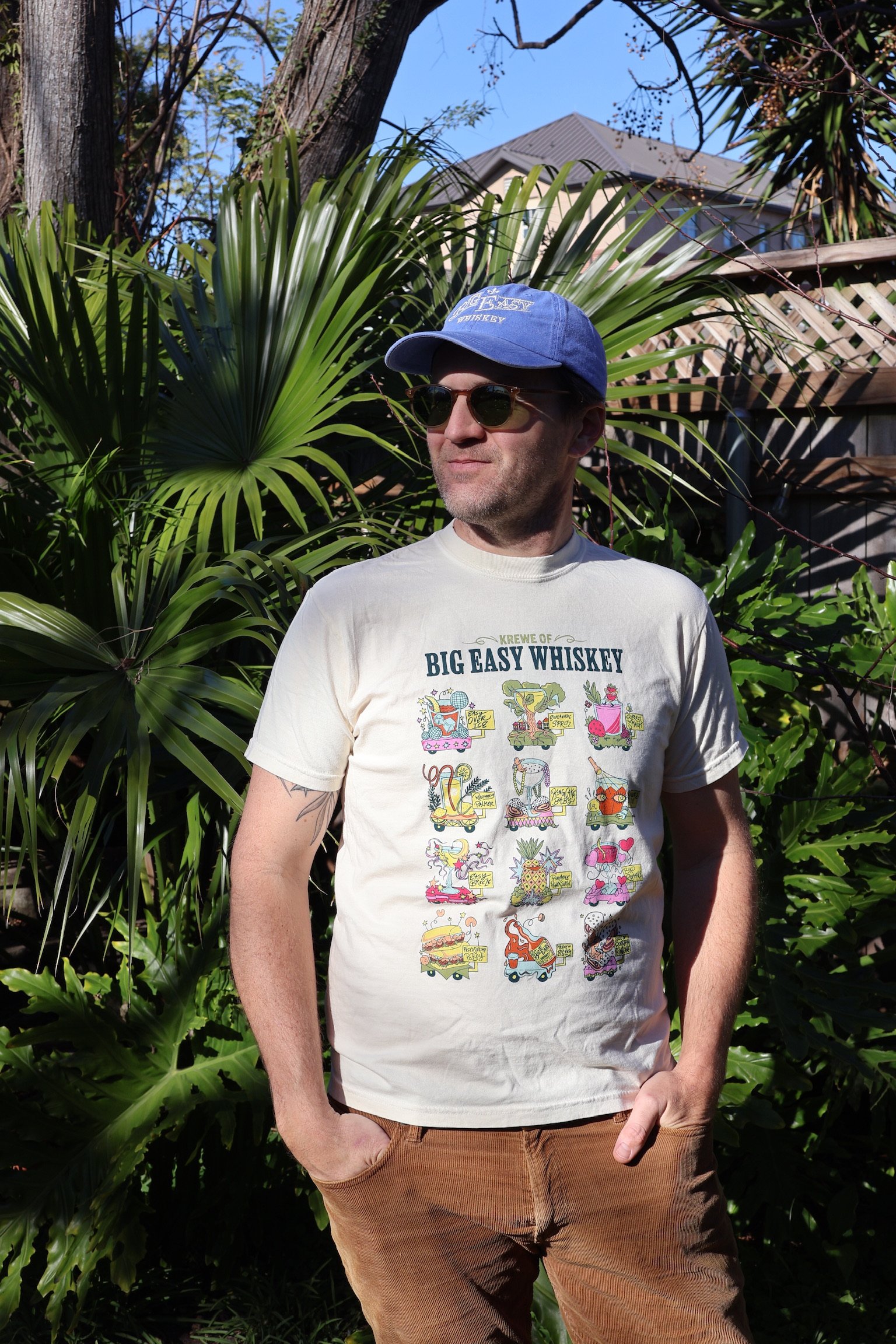 A man in sunglasses, a blue cap, and a Big Easy Whiskey t-shirt stands outdoors with a background of lush green plants and a house with a sloped roof.