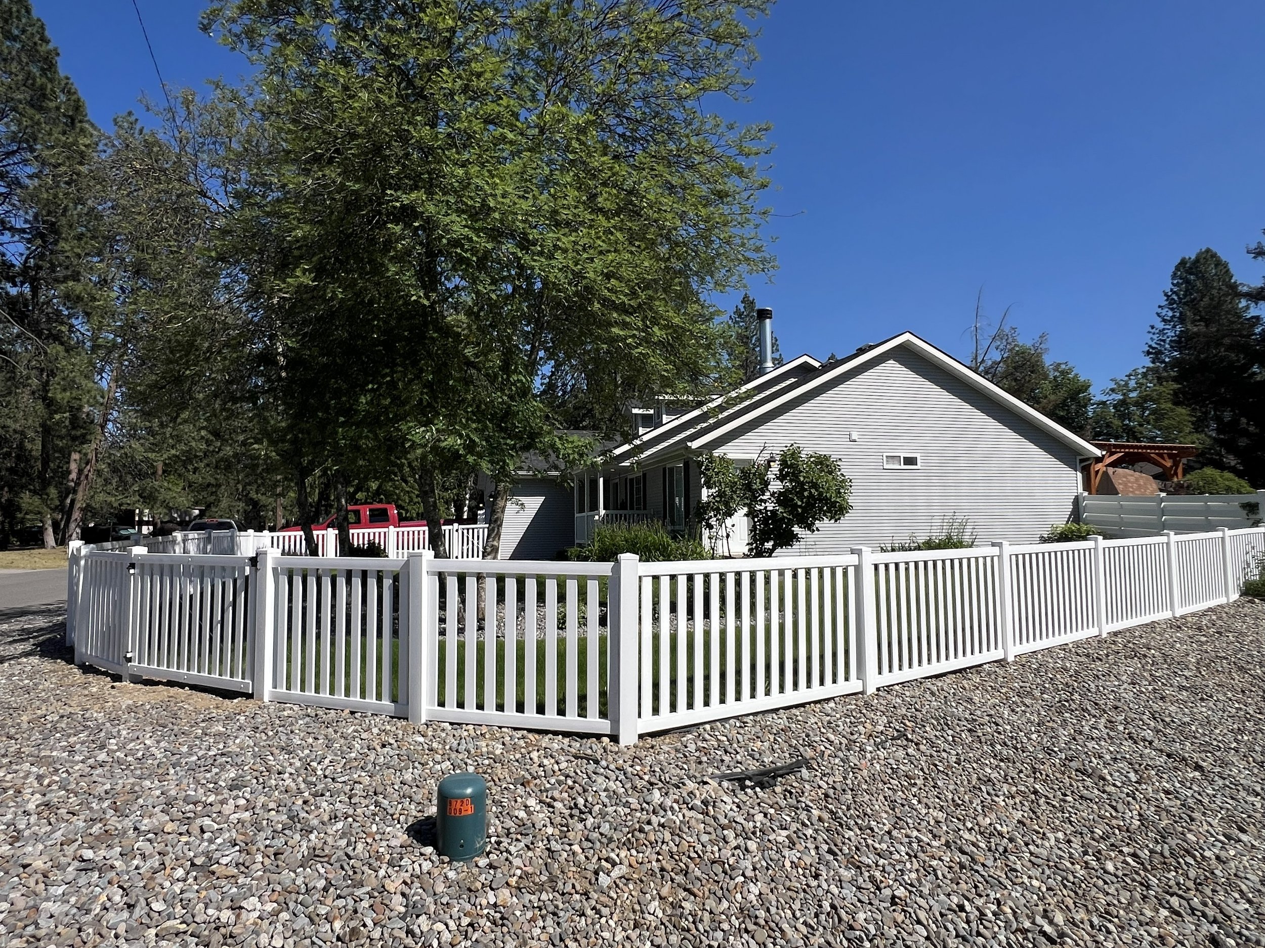 A house with light grey siding and a white picket fence around the front yard, with gravel on the ground and green trees behind the house under a clear blue sky.