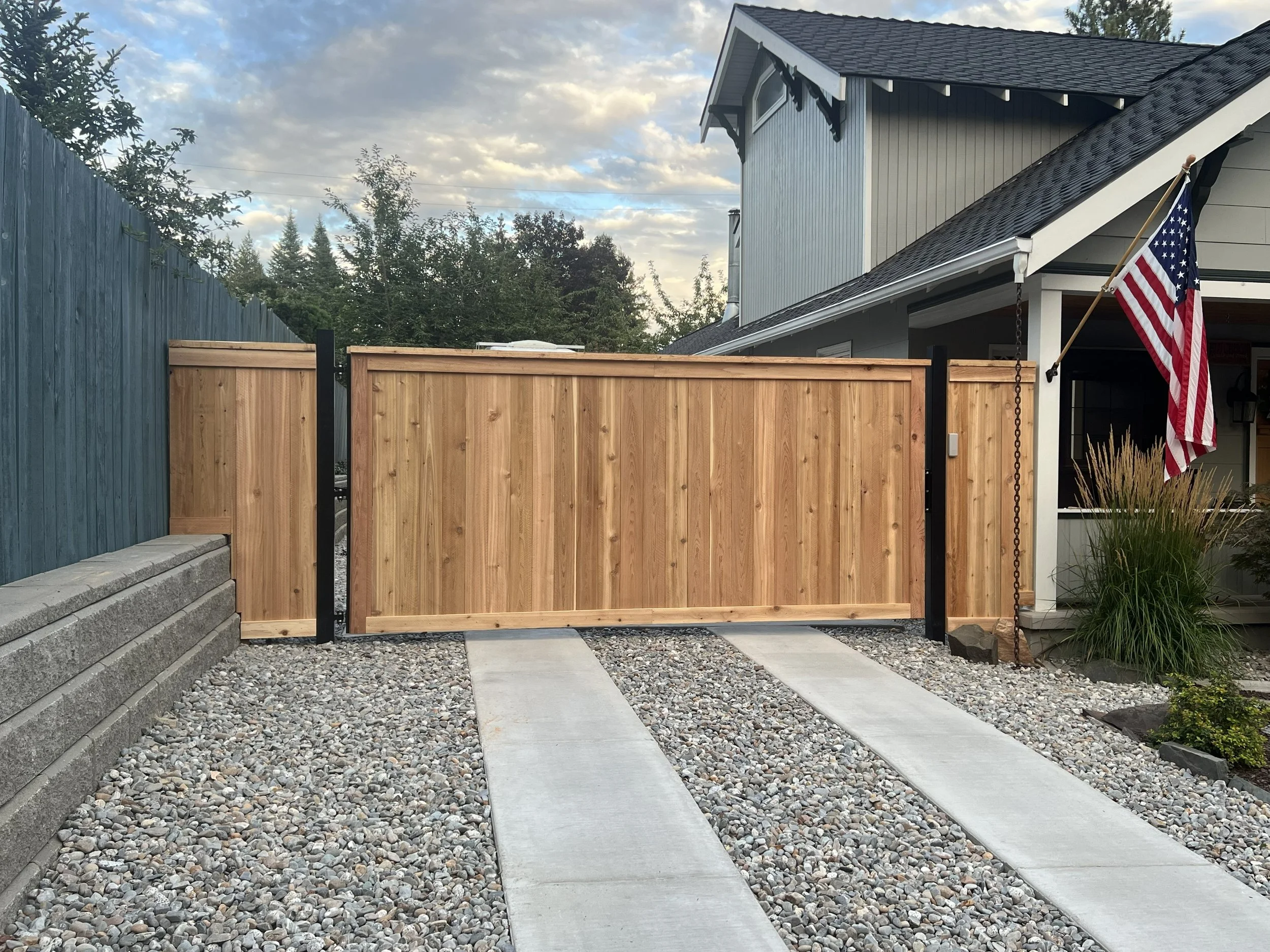 A backyard view showing a wooden privacy gate at the end of concrete and gravel driveway, with a blue fence on the left and a house with an American flag on the right.