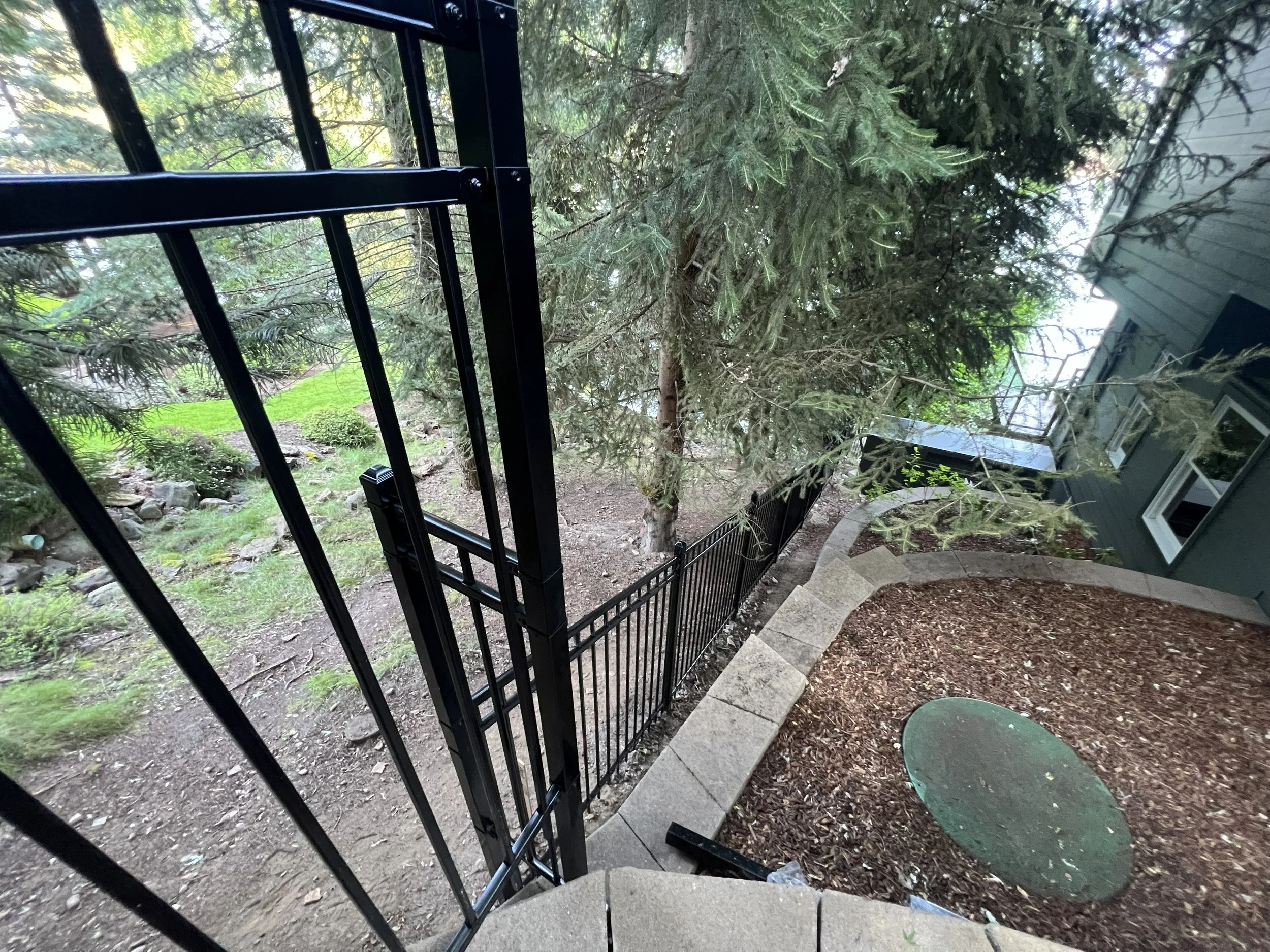 View of a backyard yard with trees, a black metal fence, and a stone pathway leading to a house, captured from the top of a staircase.