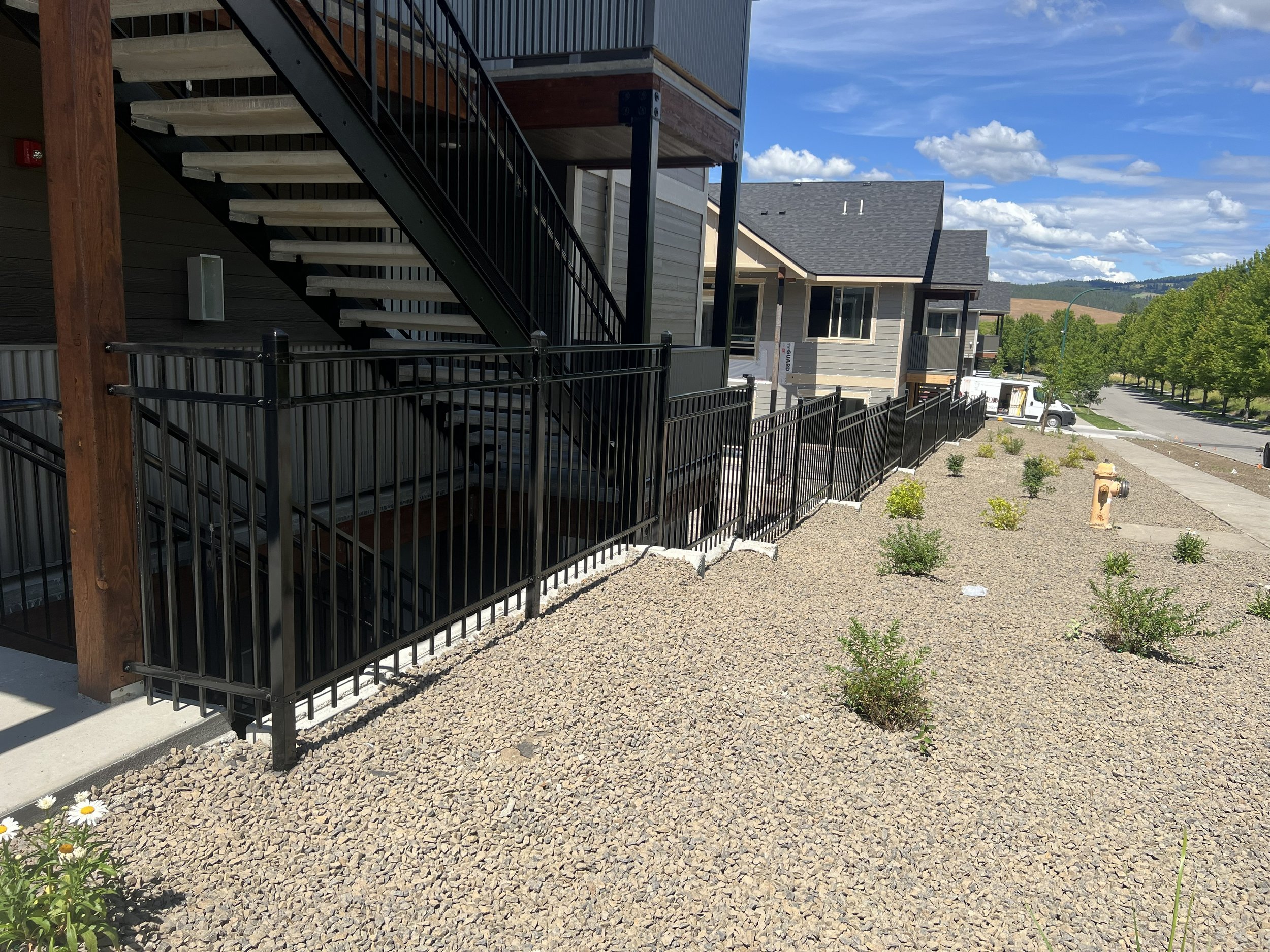 Modern residential apartment complex with exterior staircase, black metal fencing, and small landscaped yard with young plants and gravel ground cover under a blue sky with scattered clouds.