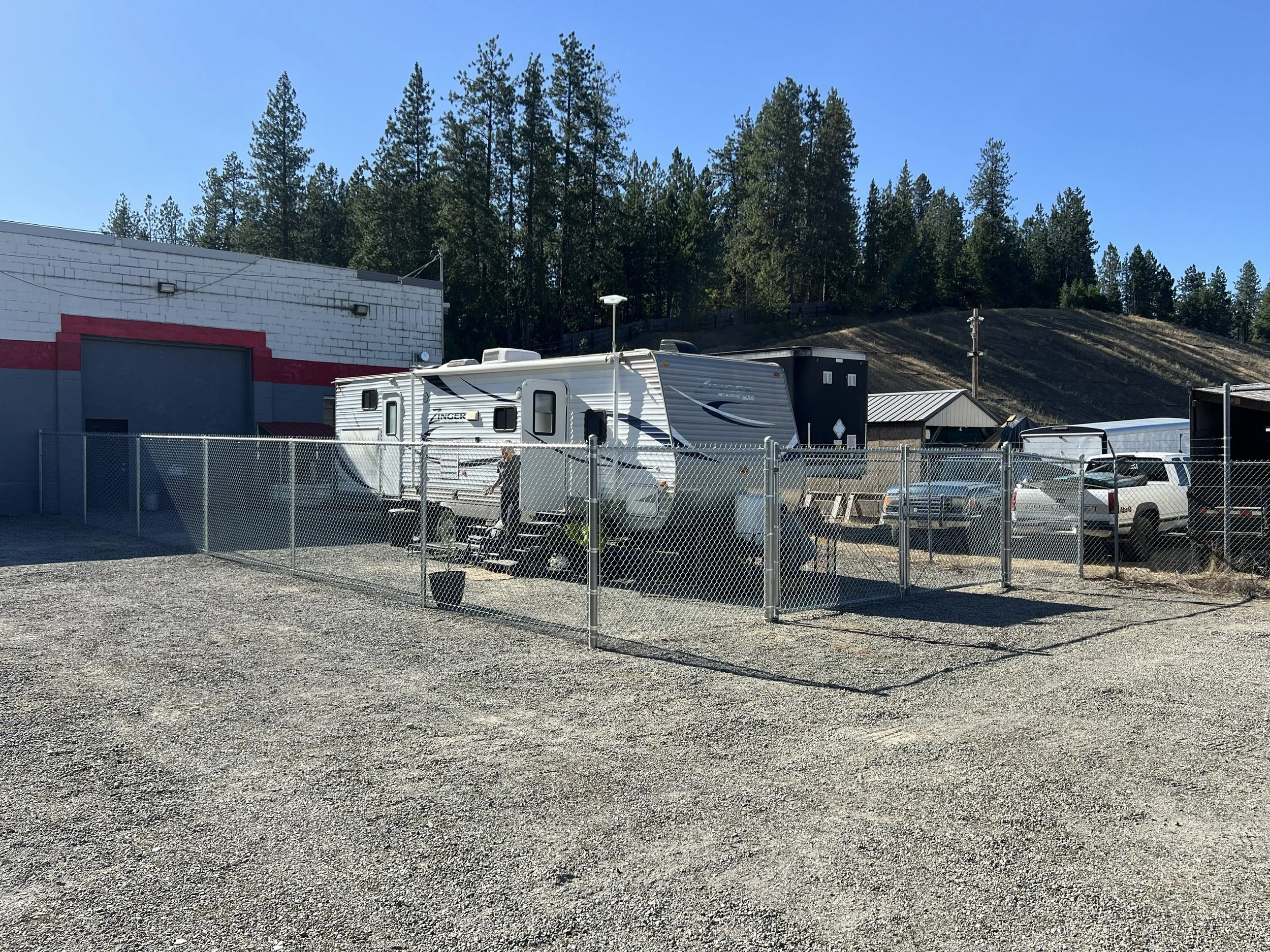 A fenced parking lot with a white travel trailer, several vehicles, and a warehouse building with a red and gray facade. In the background, there are trees and a hillside.
