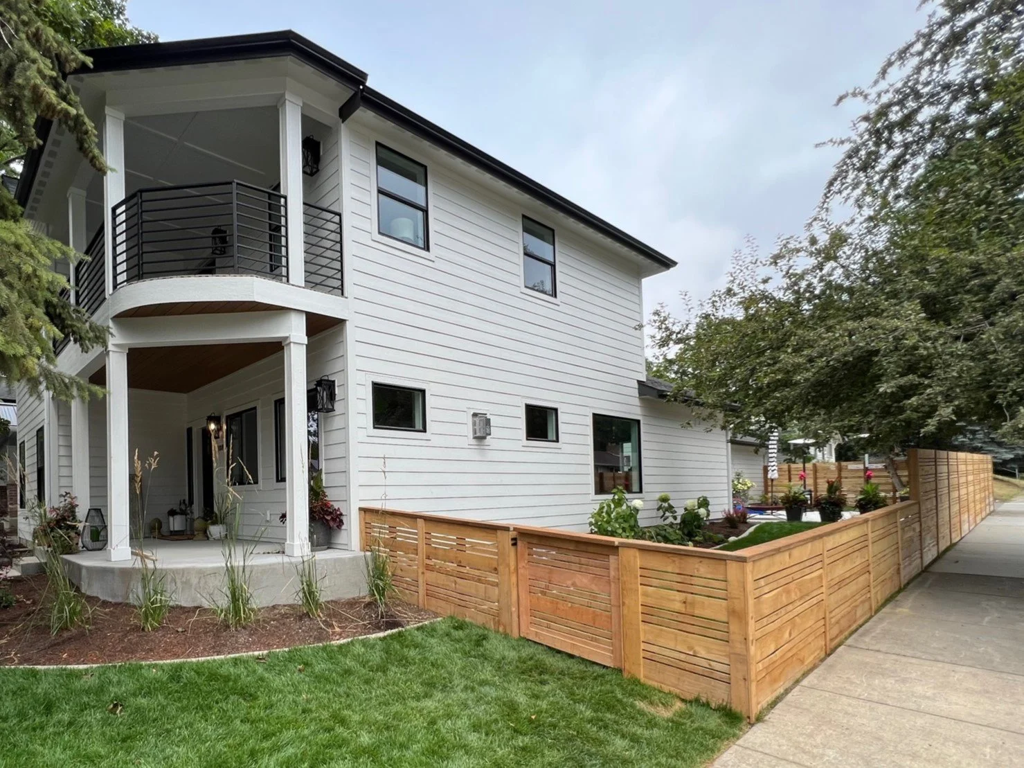 A modern white house with black trim and a second-story balcony, surrounded by a wooden fence and a green lawn with landscaping plants.