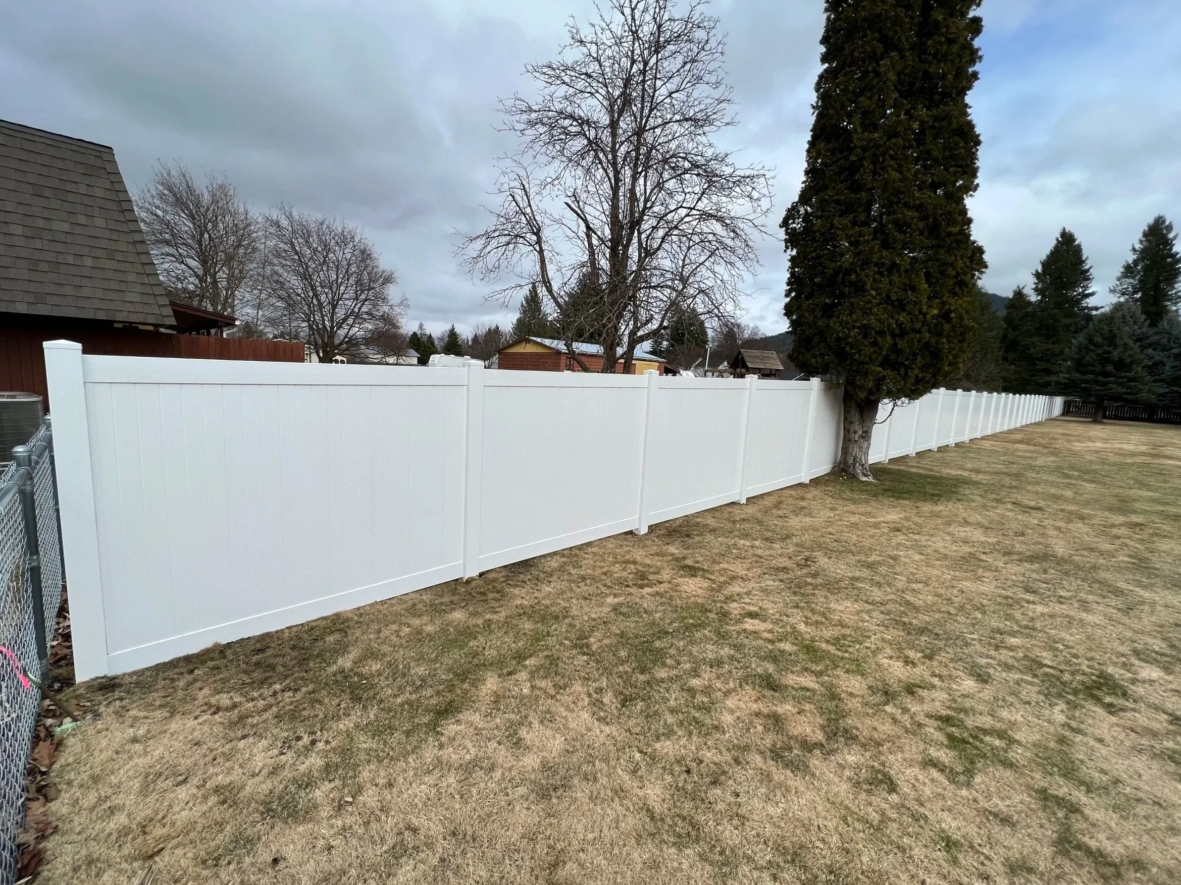 A white privacy fence in a backyard with grass and trees, overcast sky.