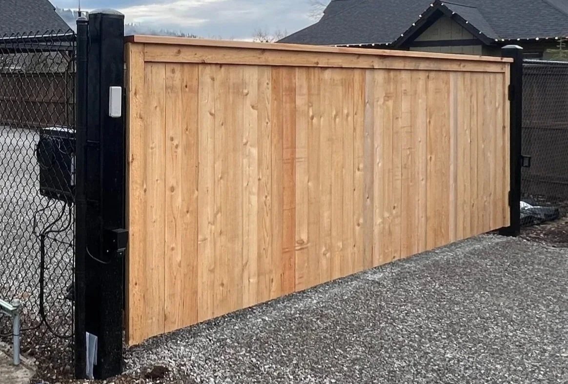 A wooden sliding gate installed between a black metal fence and a gravel ground in a residential neighborhood.