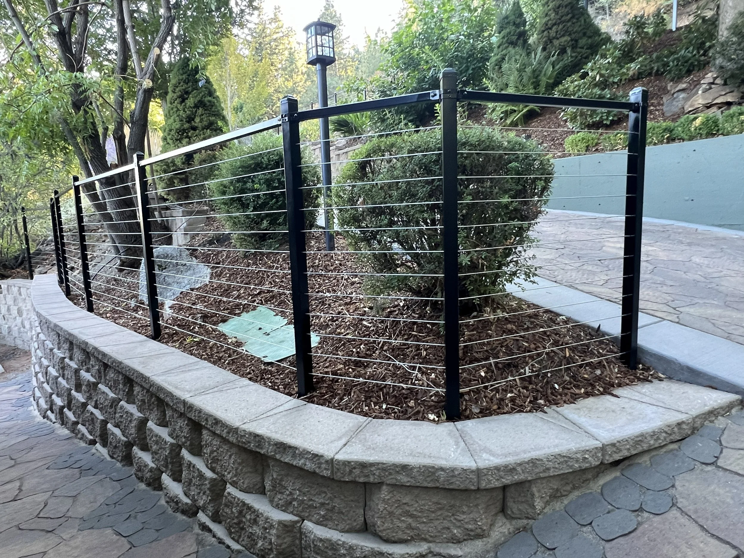A garden bed made of stone blocks with soil and shrubbery inside, enclosed by a black metal fence with horizontal wires, located along a stone pathway.