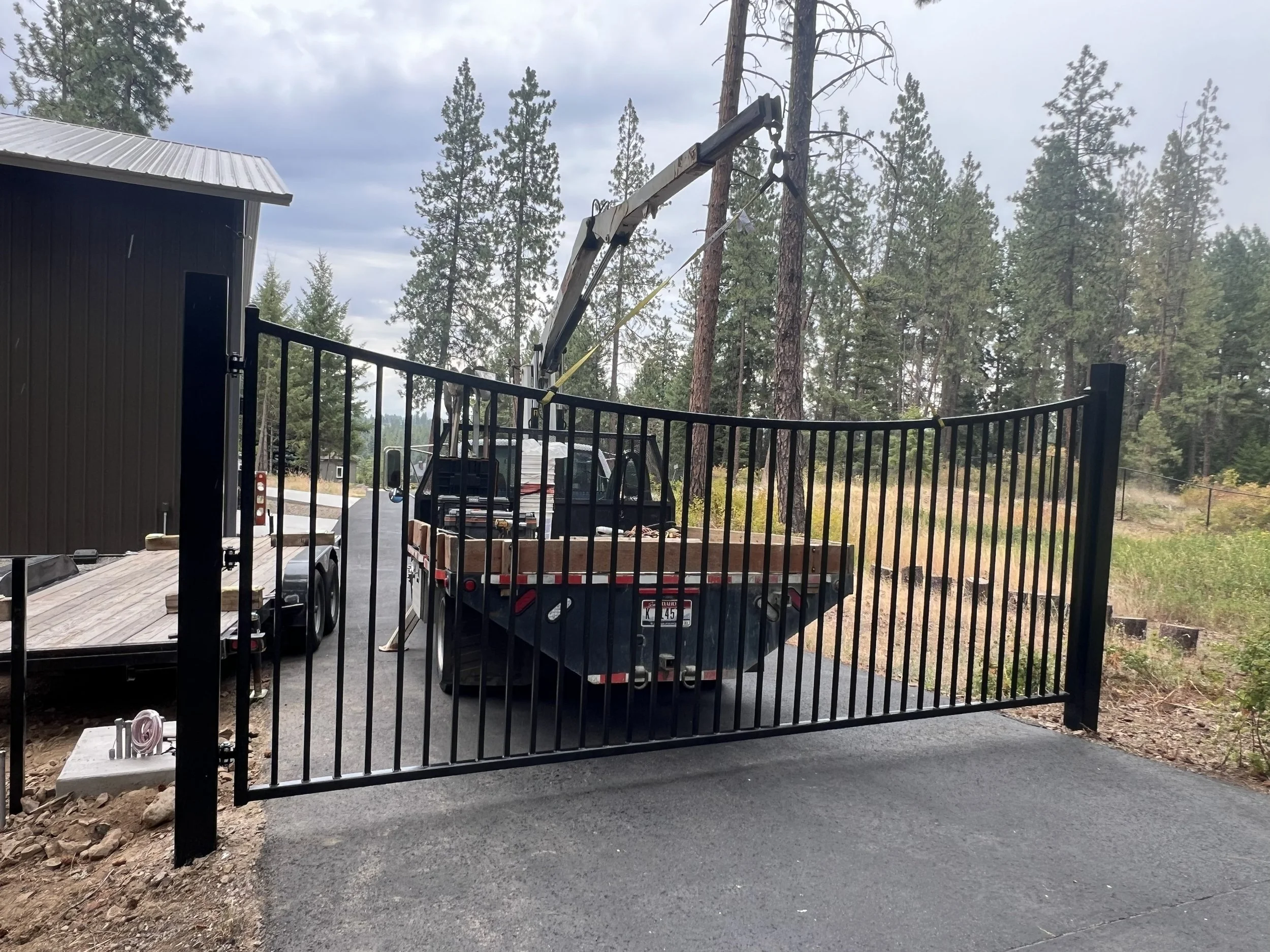 A black security gate with vertical bars in front of a driveway, with a trailer and a truck equipped with a crane in a wooded outdoor area.