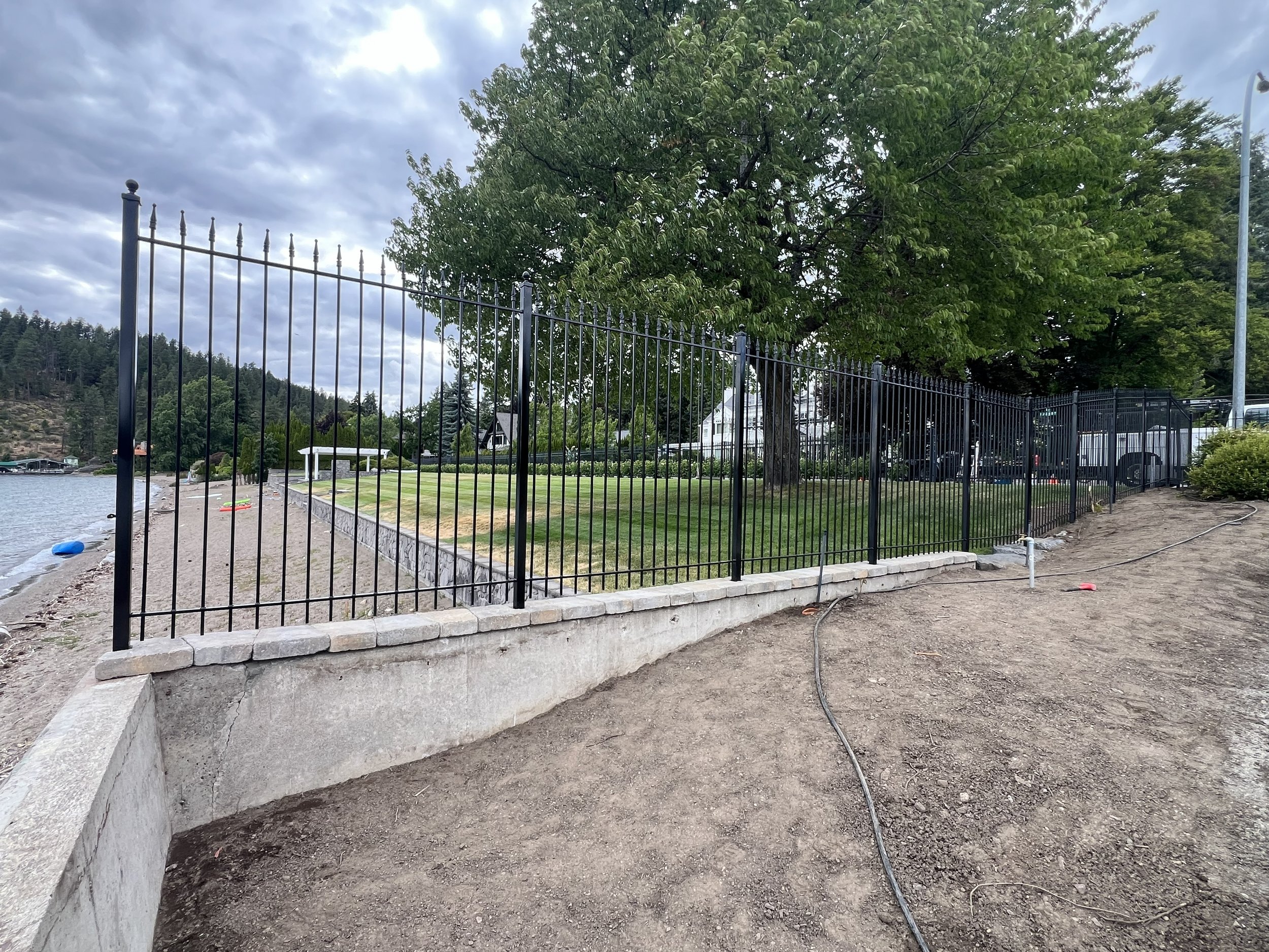 Black metal fence installed along a shoreline with trees and houses in the background, partially completed with construction materials and dirt visible.