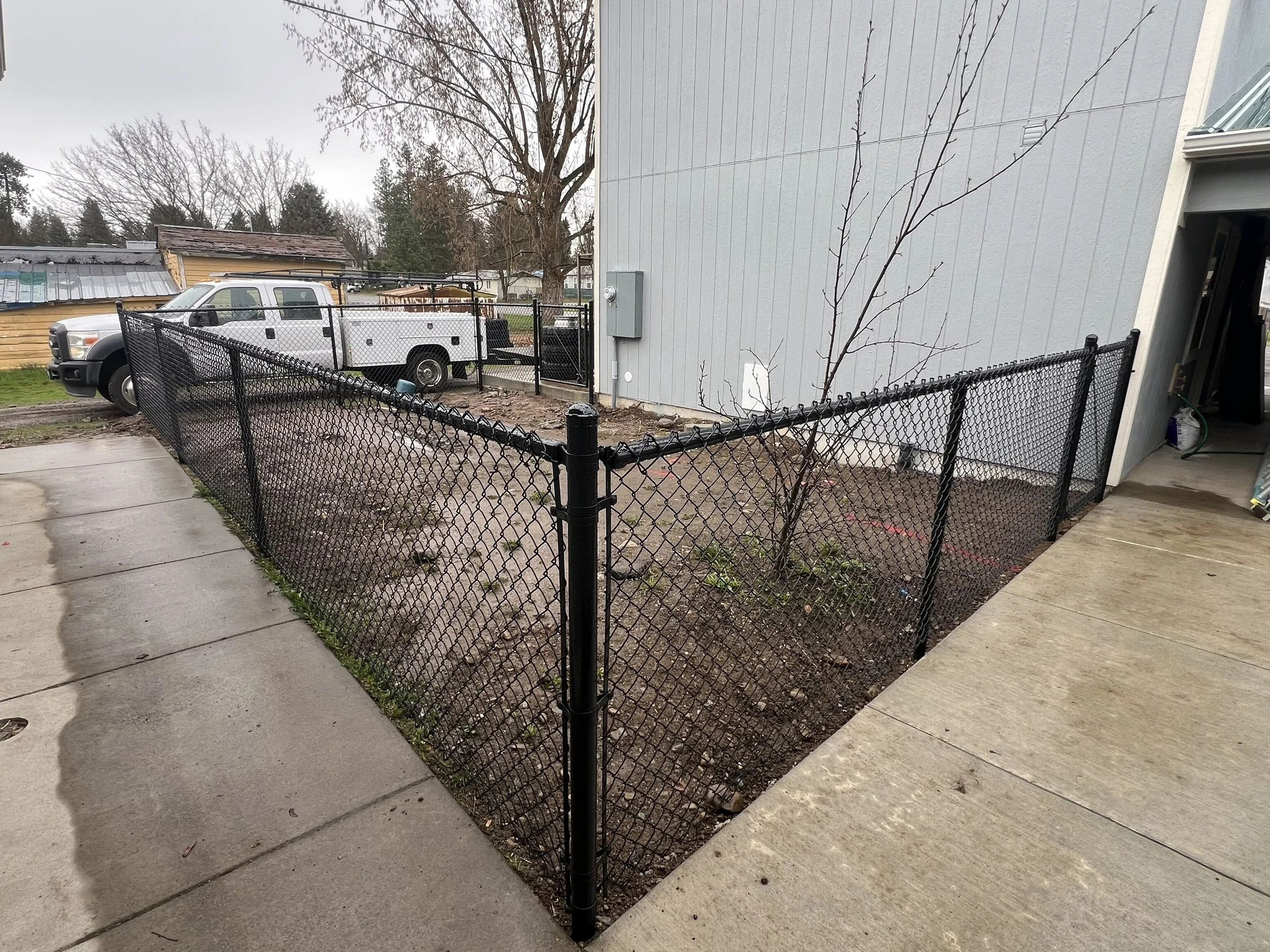 Black chain-link fence surrounding a small dirt yard next to a white house with a gray exterior. A young bare tree is inside the yard. In the background, there is a white truck and a chain-link gate.