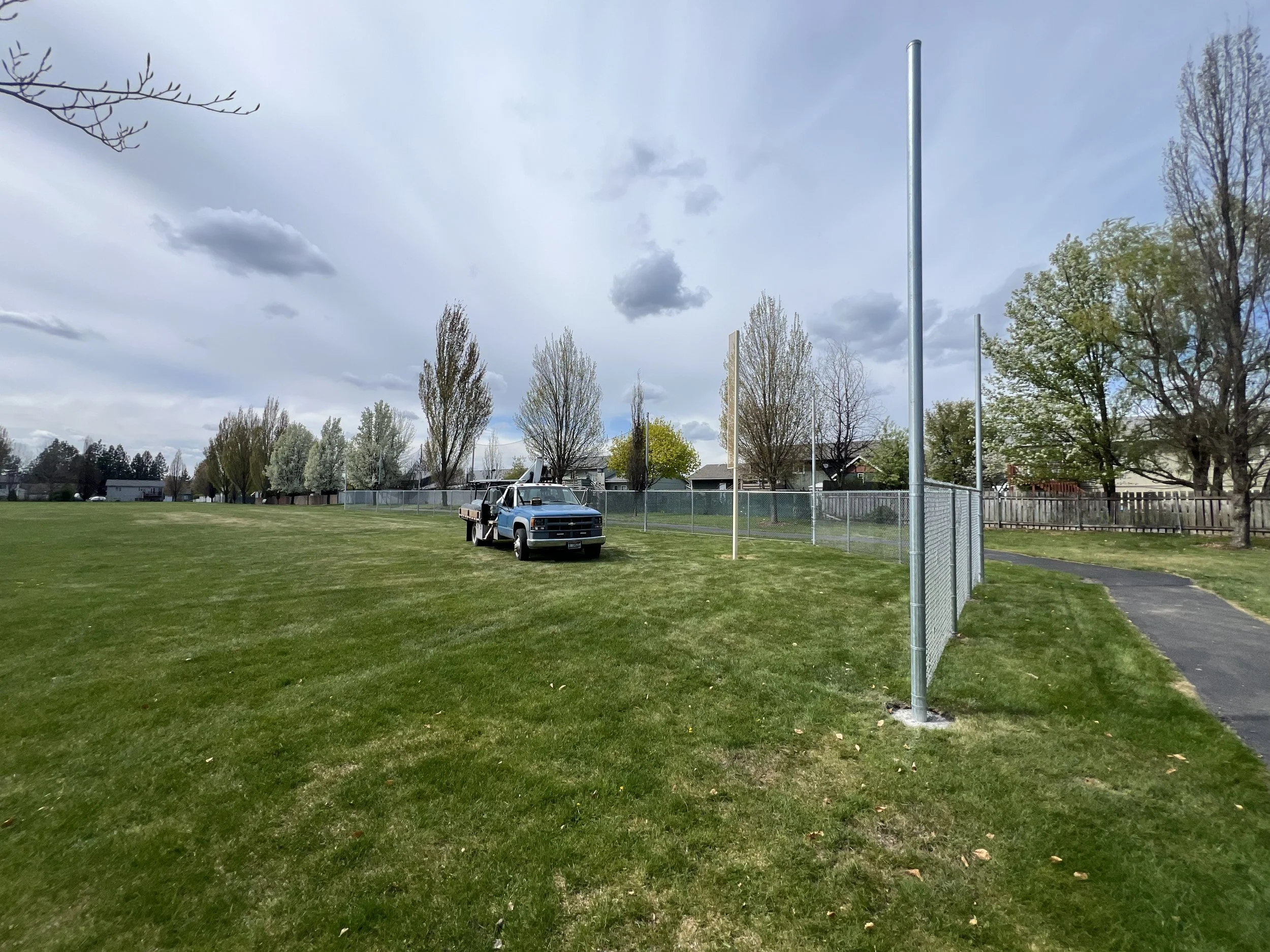 A grassy sports field with a fence, trees, a pickup truck parked near the fence, and a cloudy sky overhead.