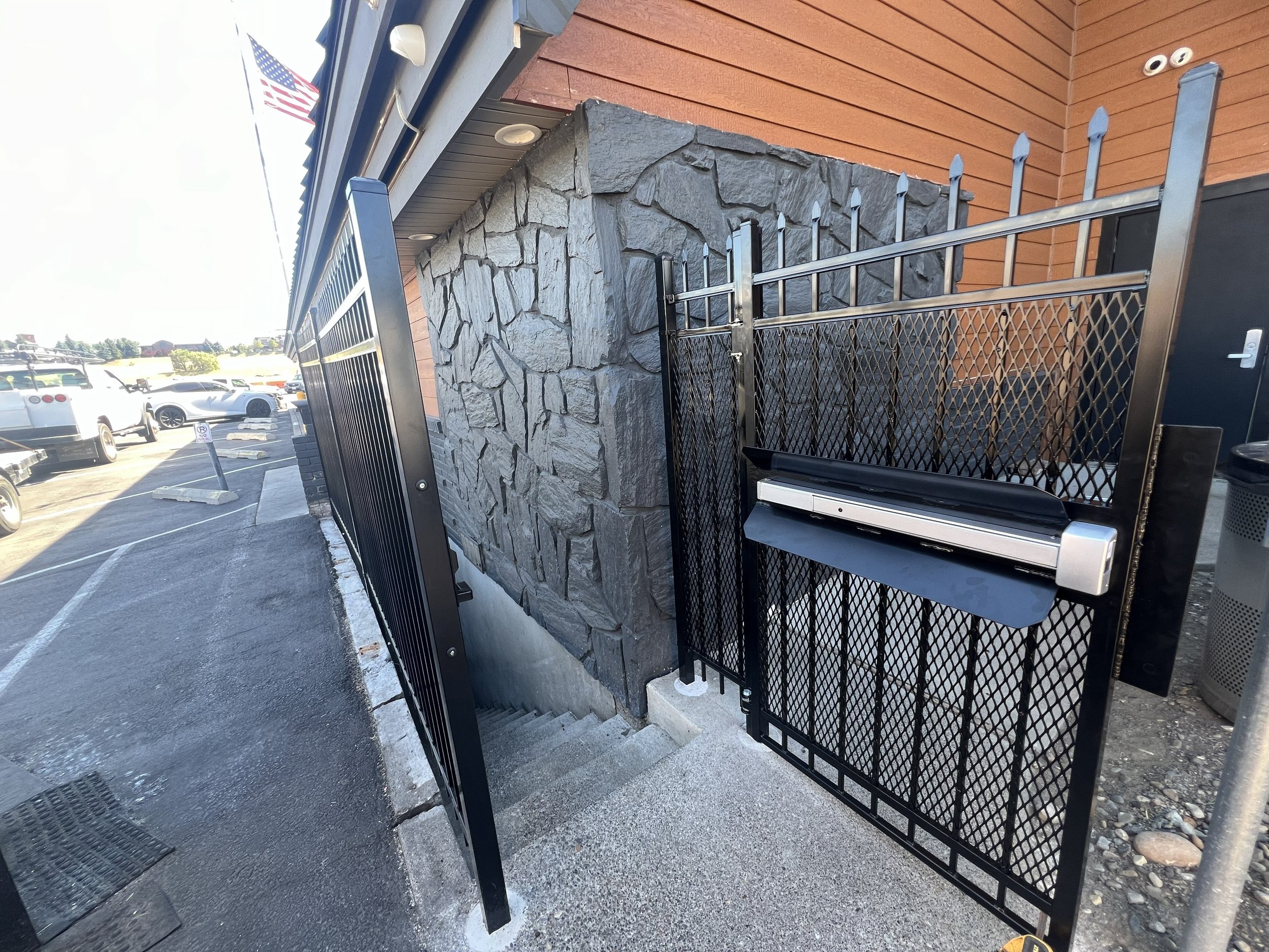 A gate with an integrated mailbox and a descending staircase leading to an underground entrance, built against a building with stone and wooden exterior walls, near a parking lot with cars.