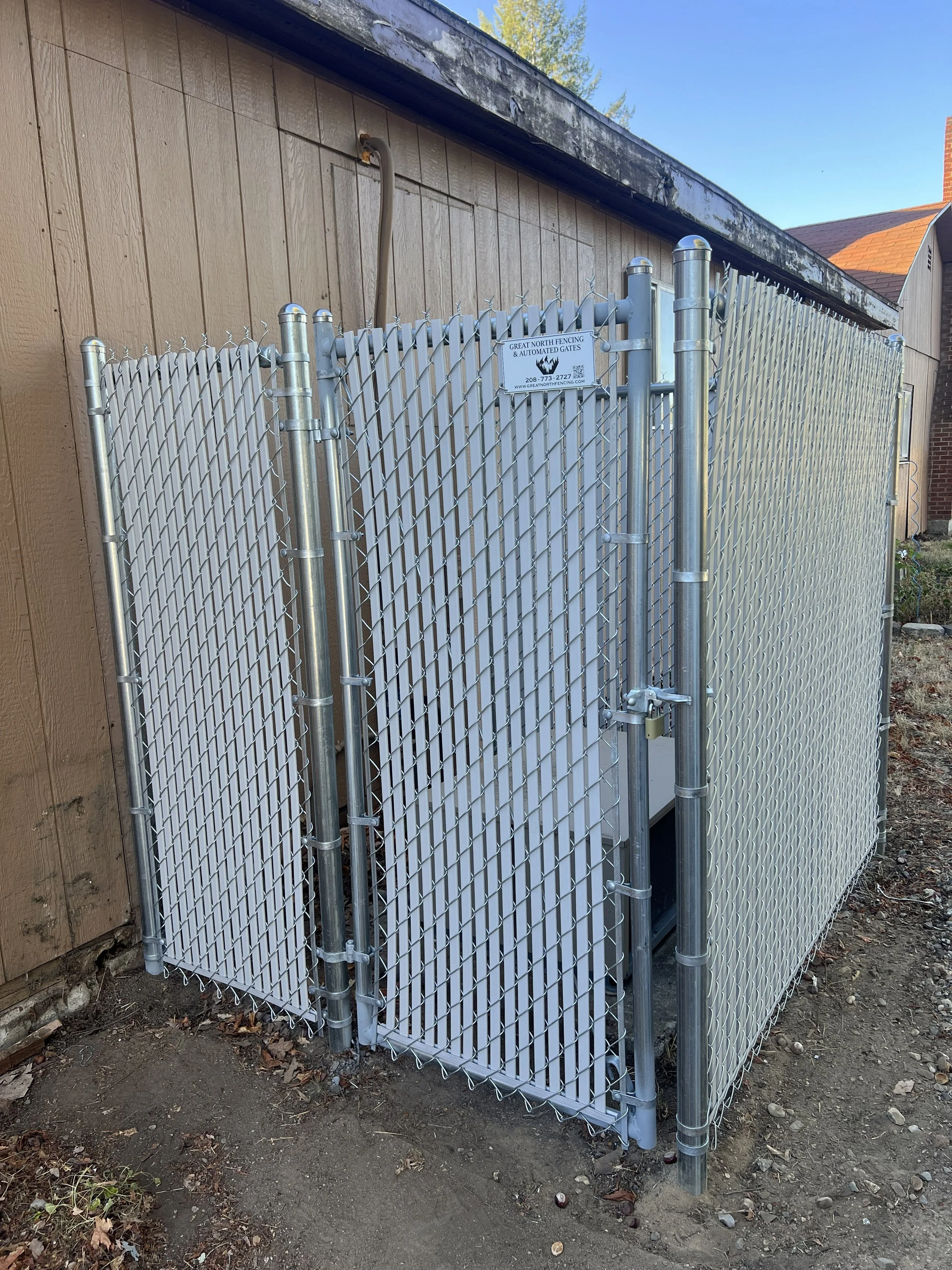 A metal chain-link fence enclosure with a gate, situated next to a wooden building, containing fencing equipment or supplies inside.