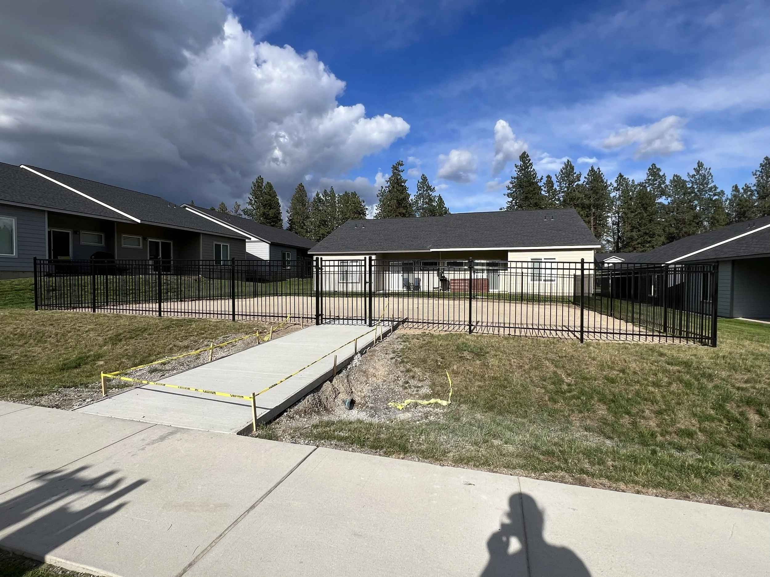 Residential backyard with a concrete pathway leading to a fenced yard, houses in the background, and partly cloudy sky.