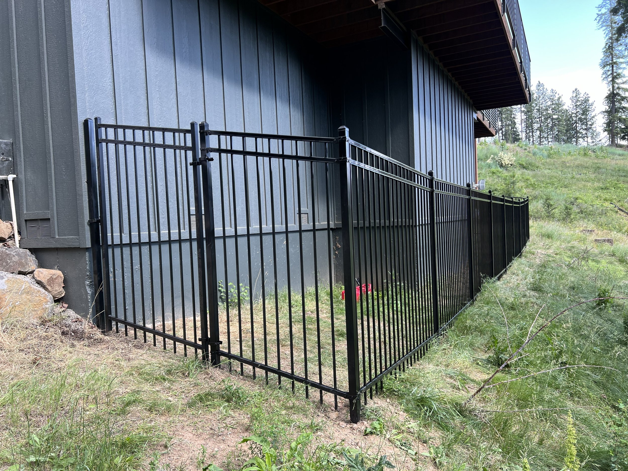 Black metal fence surrounding a small area of grass next to a blue house with vertical siding, located on a hillside with trees in the background.