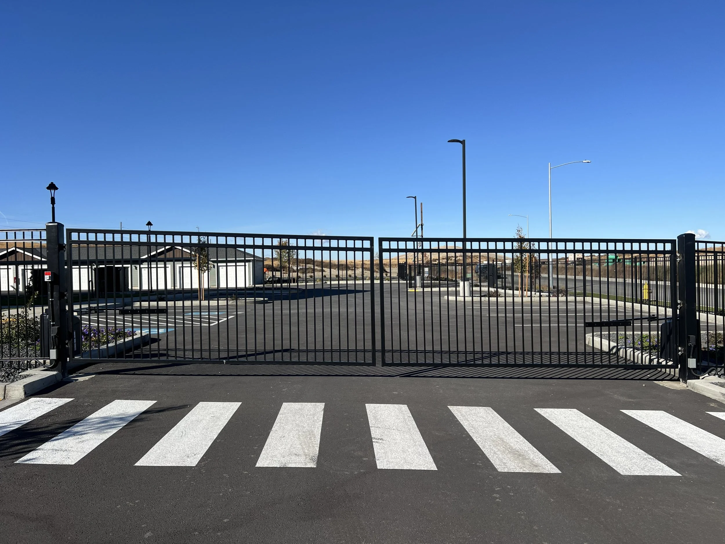 A closed black metal gate in front of a parking lot with white buildings, tall streetlights, and a clear blue sky in the background.