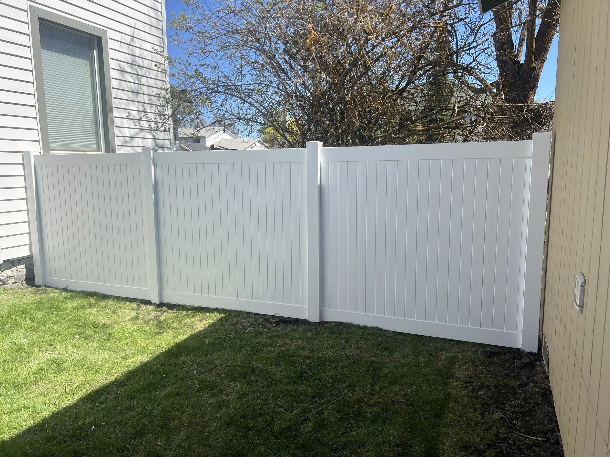 White vinyl privacy fence in a backyard, with a green lawn and a tree in the background.