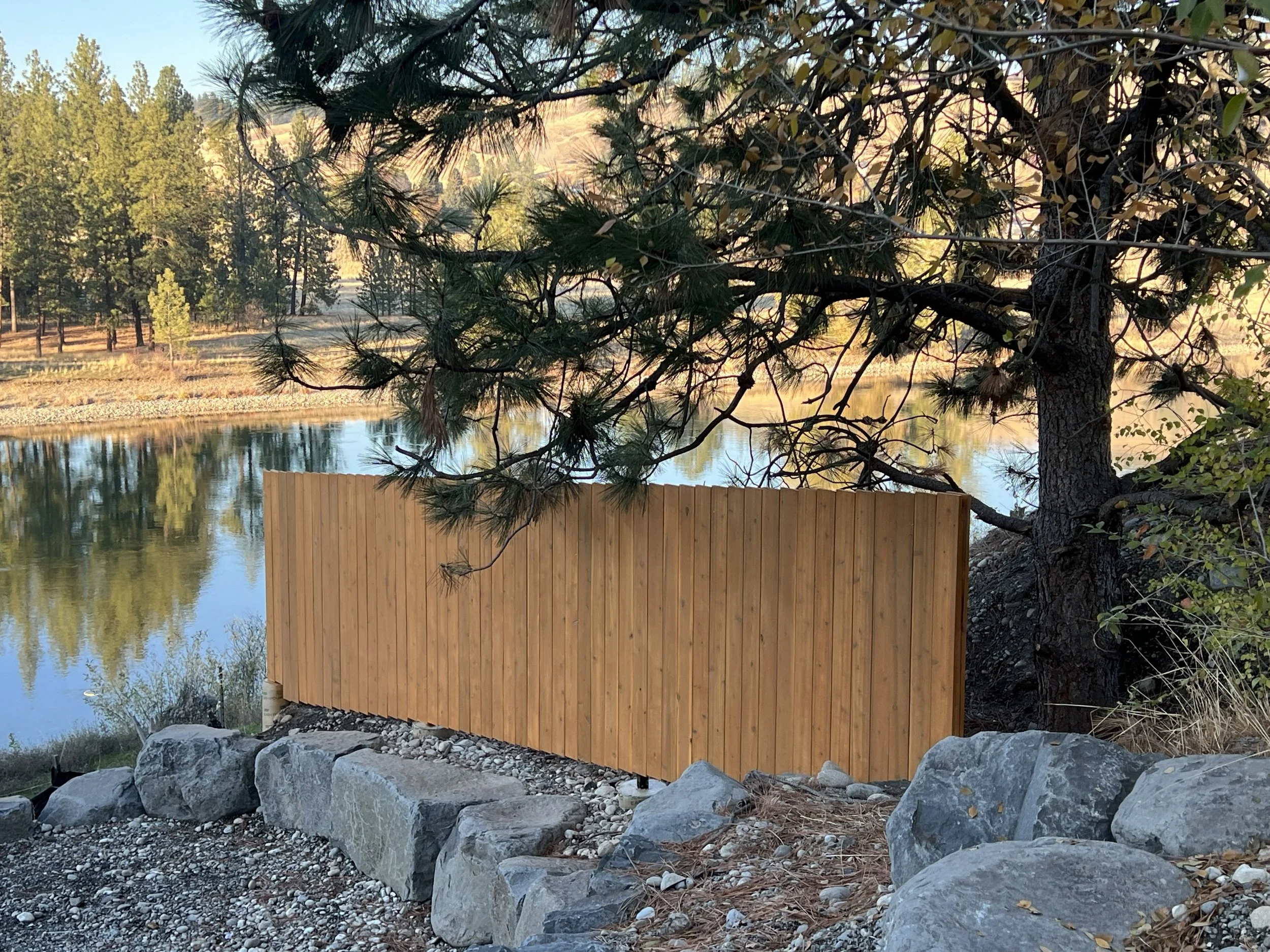 A wooden fence next to a river with rocky shoreline, surrounded by trees with a forest and hills in the background.