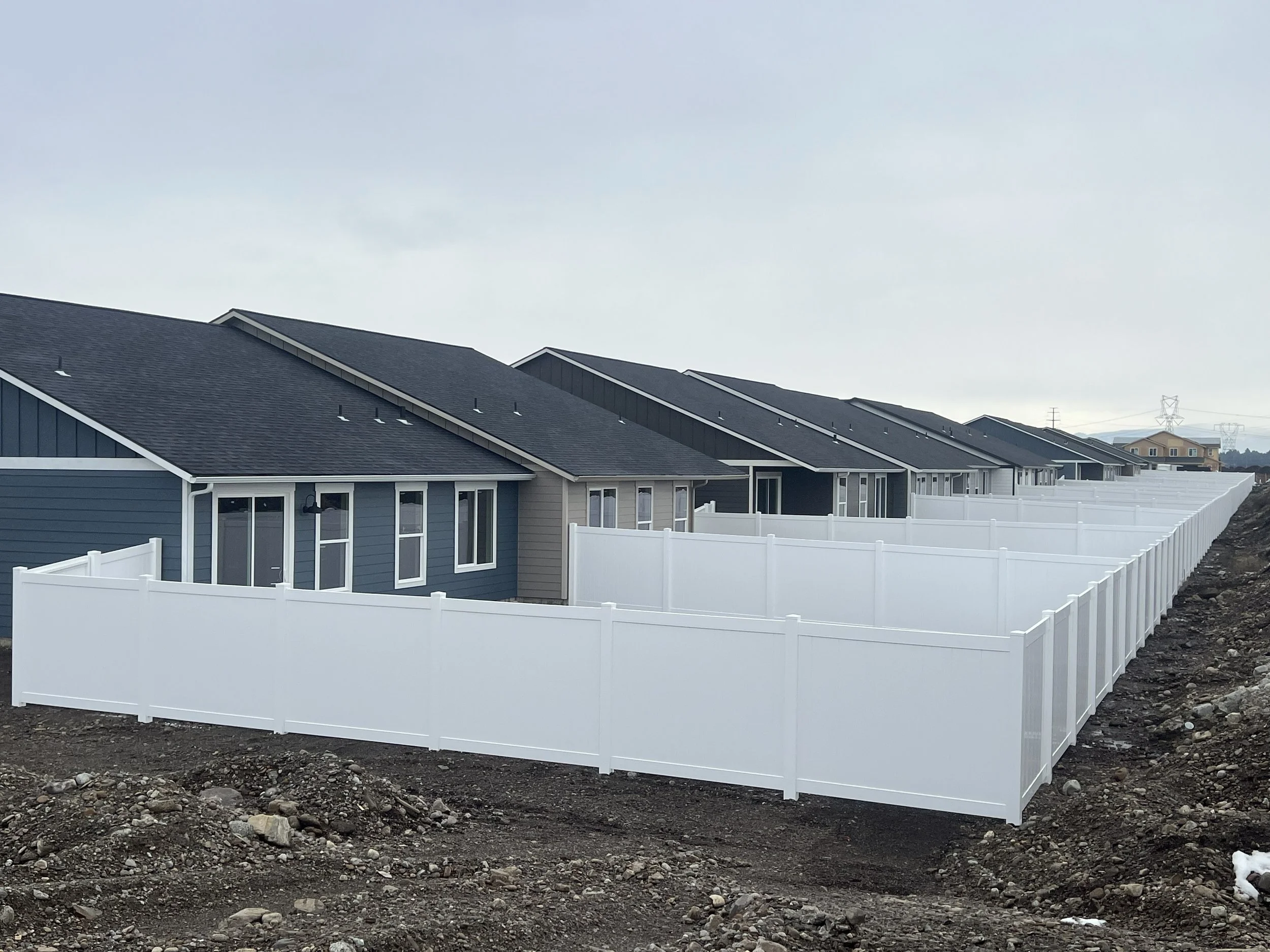 Row of new houses with dark gray roofs and blue siding, surrounded by white fencing, under cloudy sky.