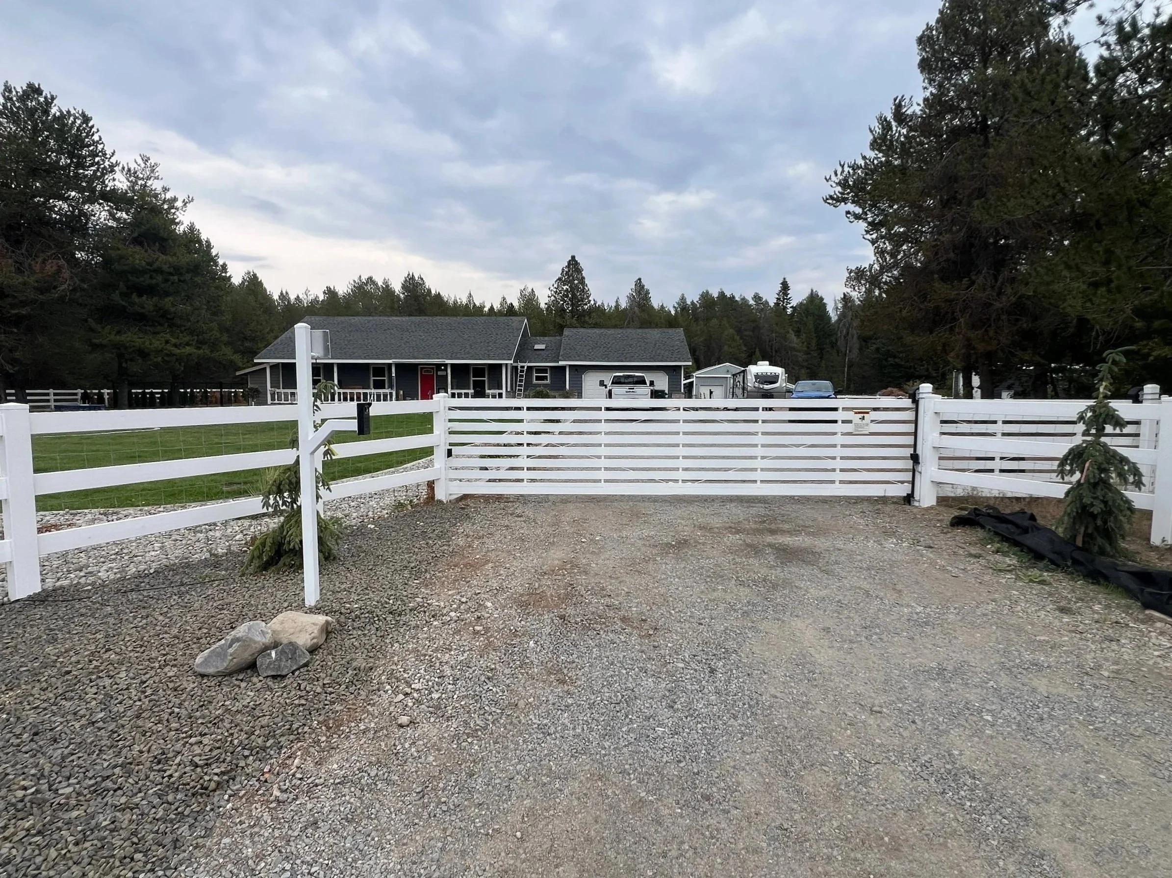 A gravel driveway leading to a white fence gate, with a house, trees, and parked vehicles in the background.