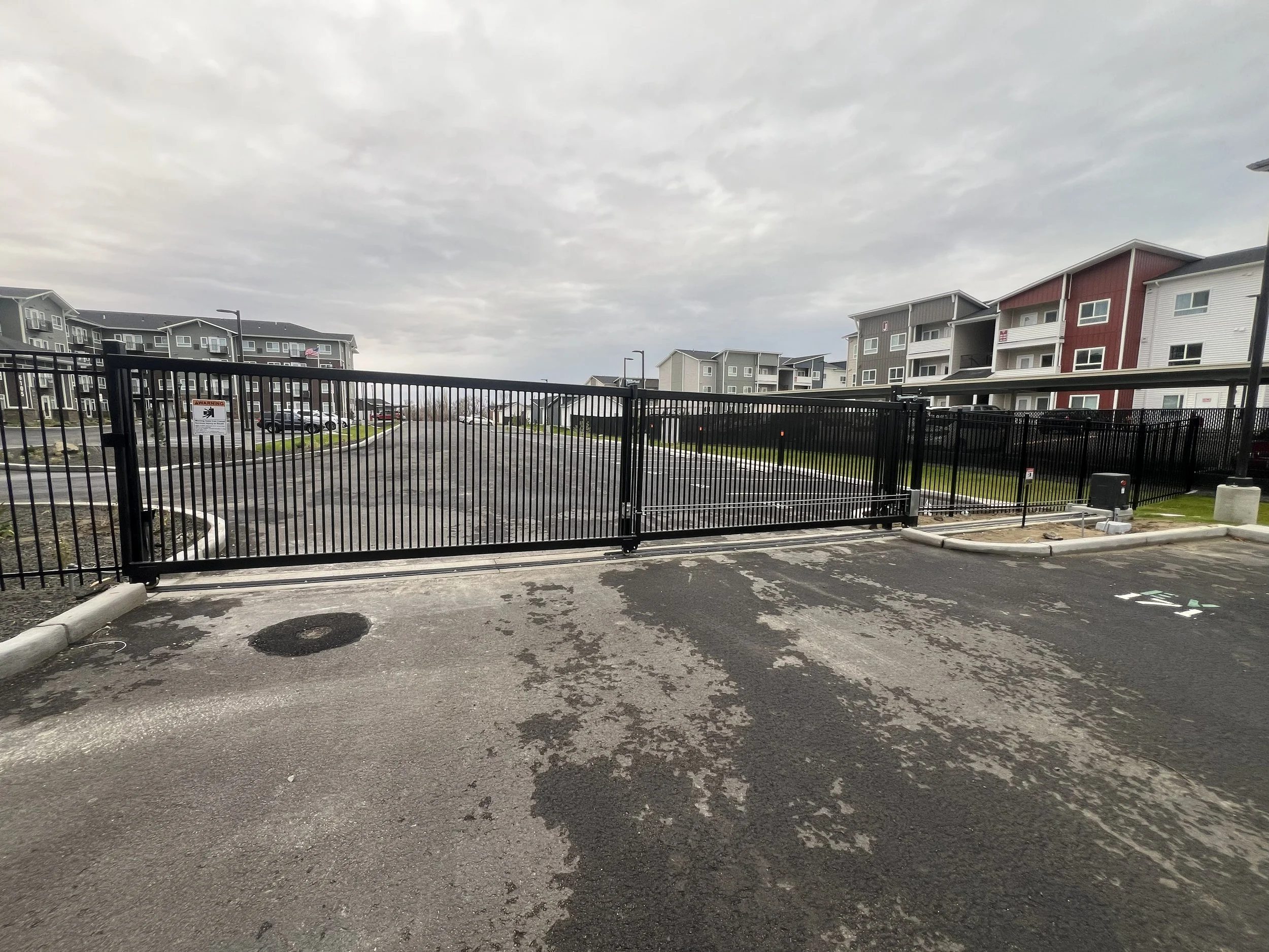 A gated entrance to an apartment complex with multi-story buildings in the background, a parking lot, and overcast sky.