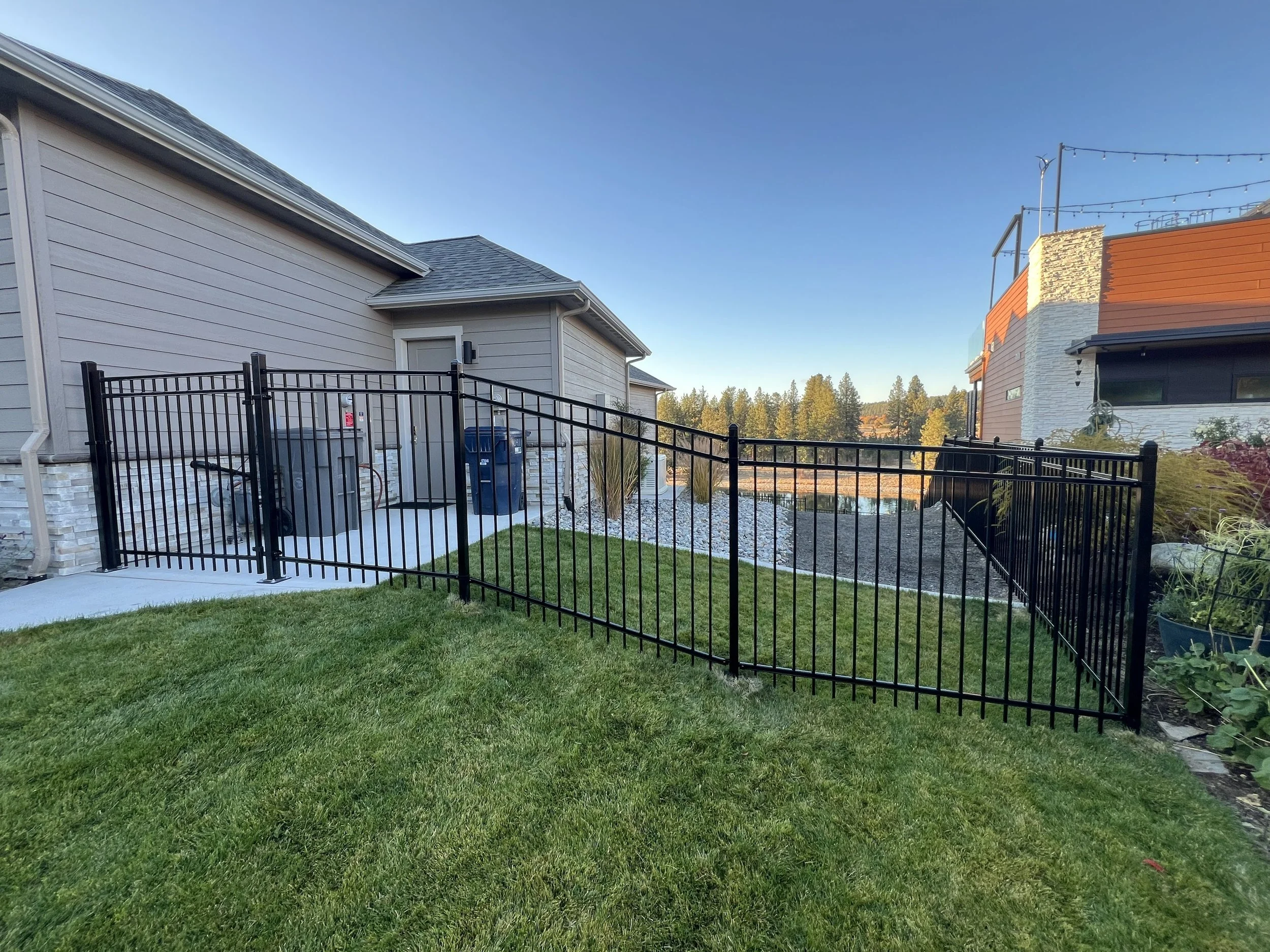 A black metal fence dividing a green lawn from a paved area with trash bins on the left. The background shows neighboring houses and trees under a clear blue sky.