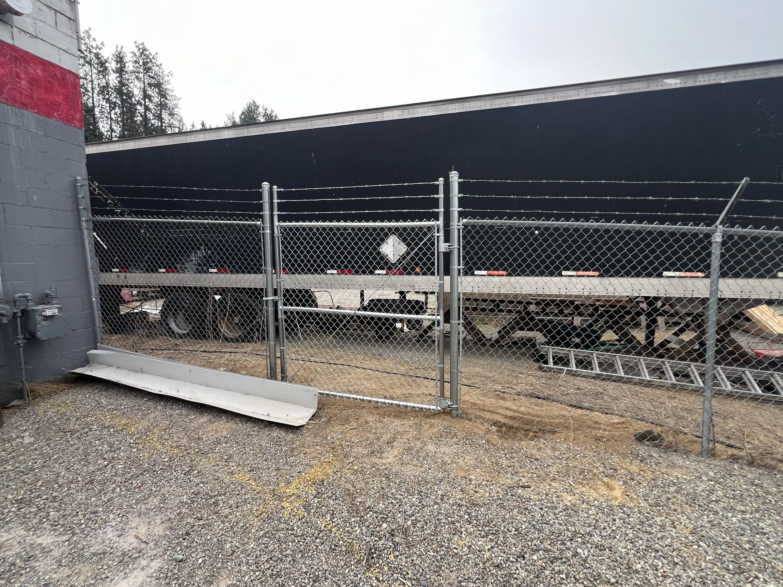 Storage yard with trailers behind a chain-link fence, with a gray building on the left and graffiti on the wall, overcast sky, surrounded by trees.