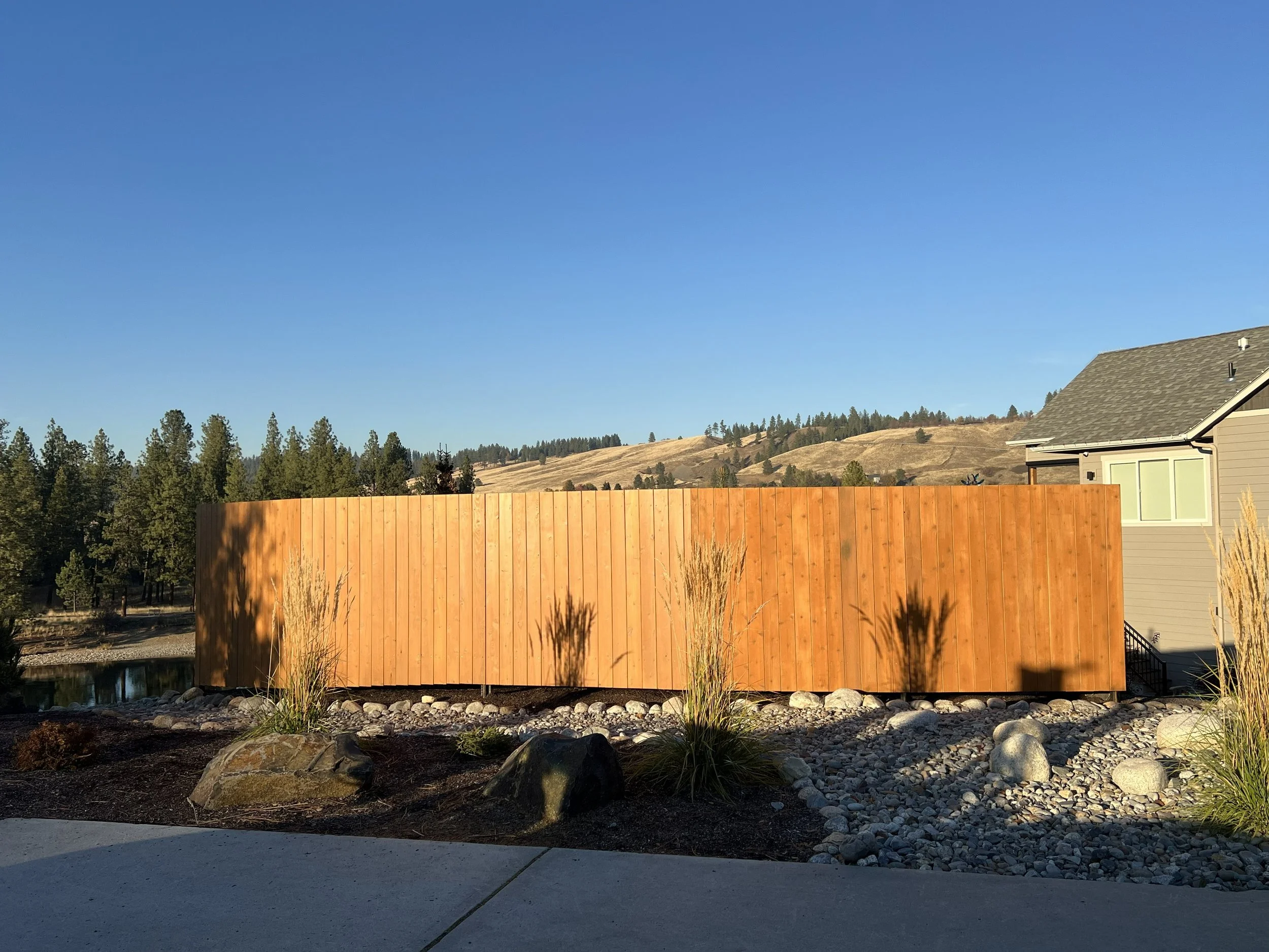 A wooden privacy fence in a backyard, with a house on the right and a landscaped area with rocks and plants in the foreground. In the background, there are trees and hills under a clear blue sky.