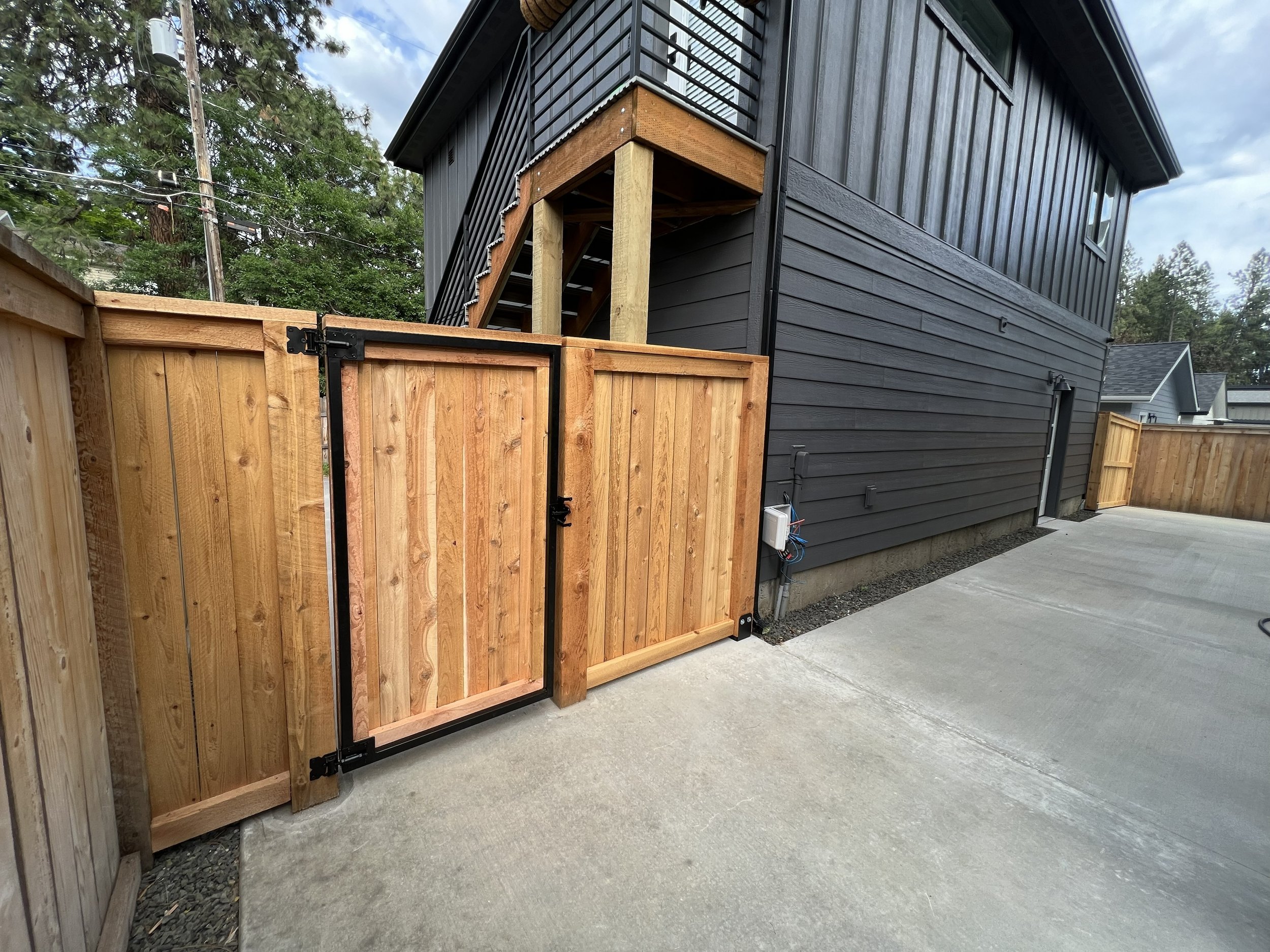 Newly installed wooden and metal gate and fence in backyard adjacent to a dark gray house with stairs leading up to a second floor. Concrete driveway in the foreground, trees in the background.