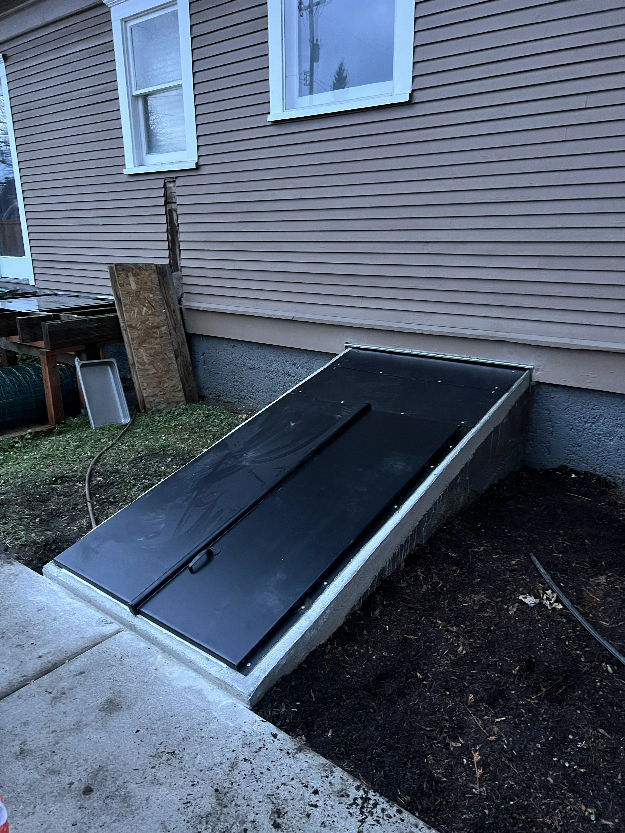 A basement window with a black metal cover installed at an angle on the exterior of a beige house. The ground surrounding the window is bare soil, and there is a patch of grass nearby.
