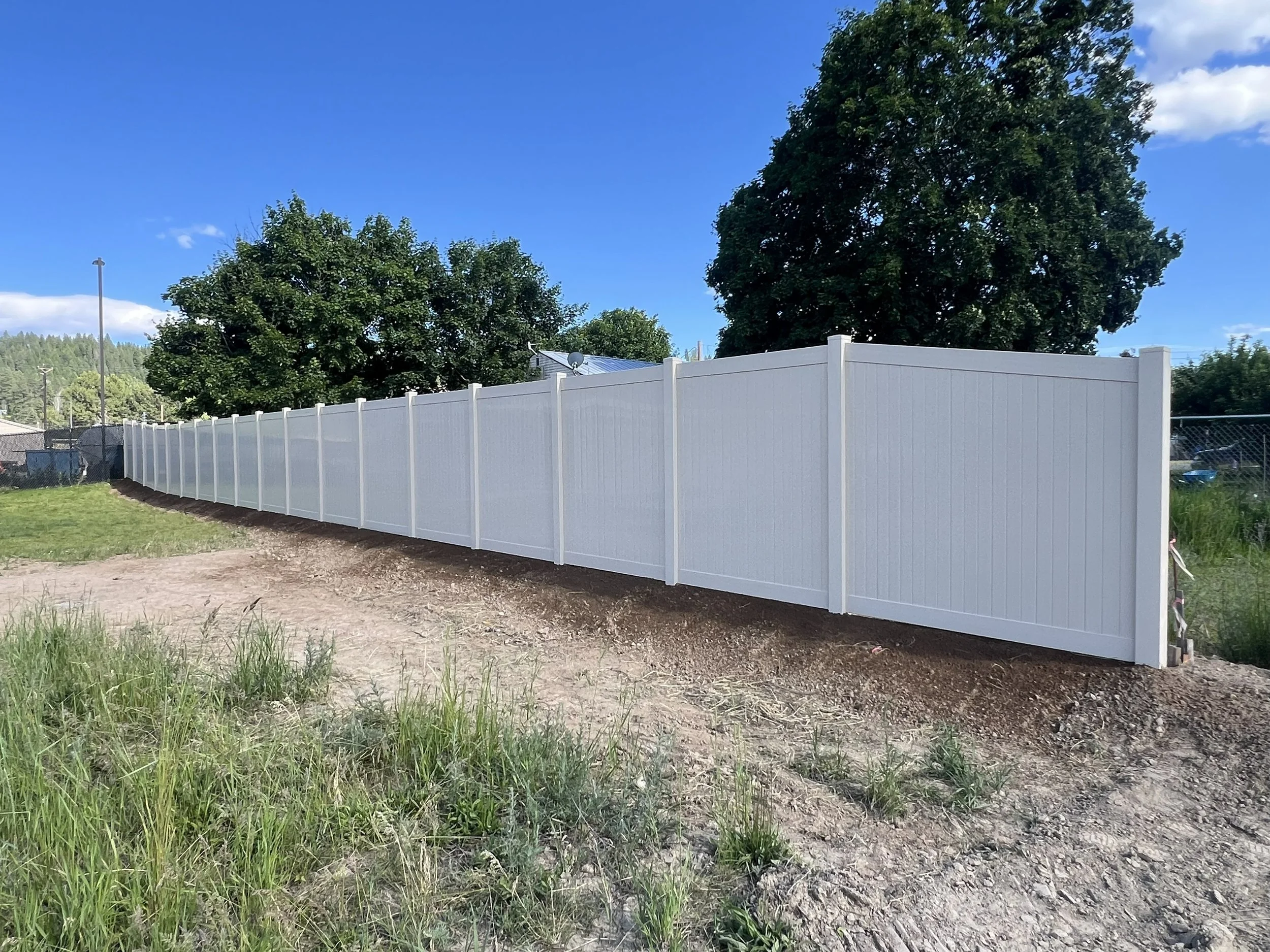A white privacy fence installed on a dirt surface under a blue sky, with green trees in the background.