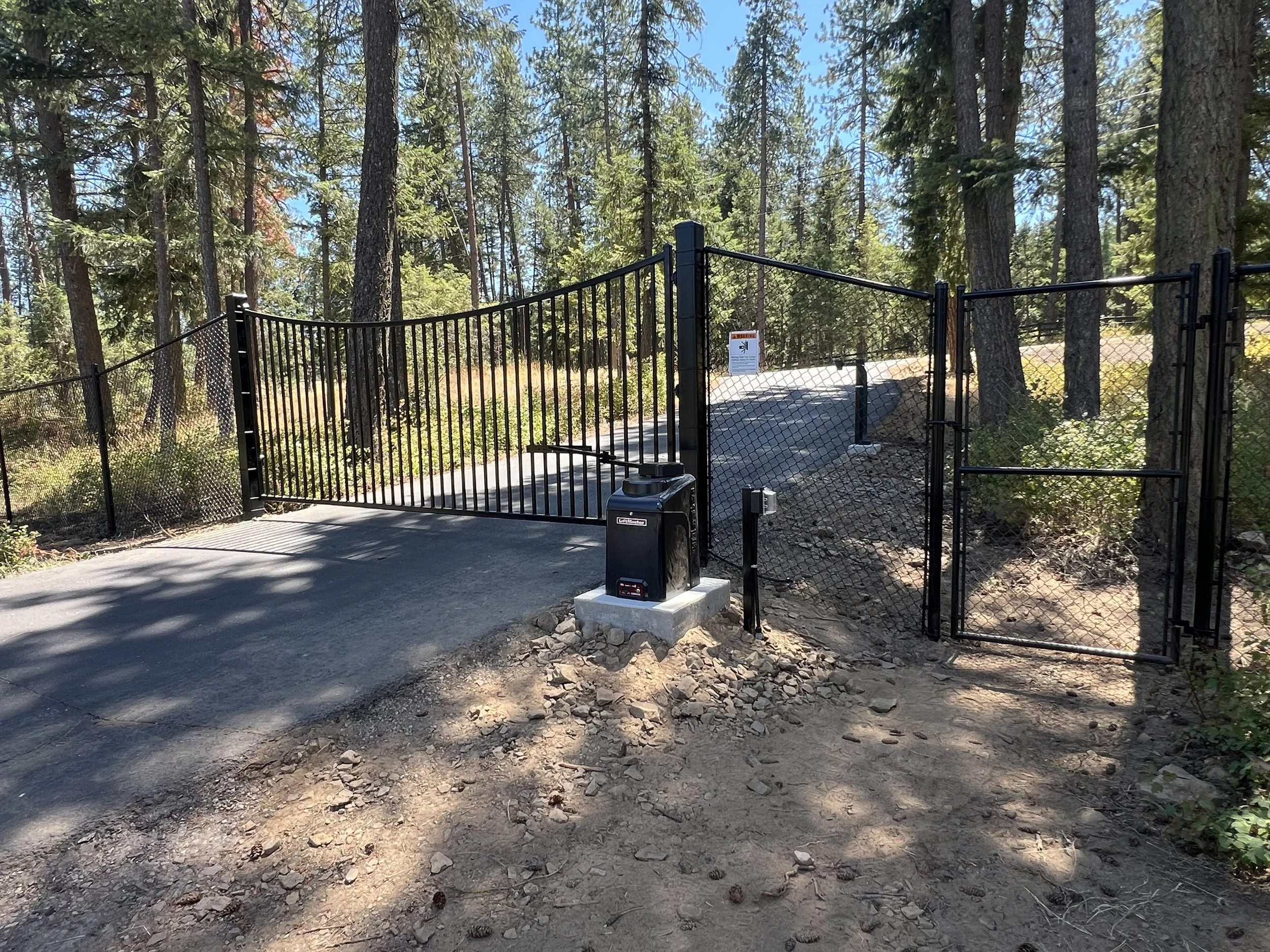 Black metal gate and fencing at the entrance of a wooded area, with a paved path beyond and surrounded by tall trees and natural vegetation.