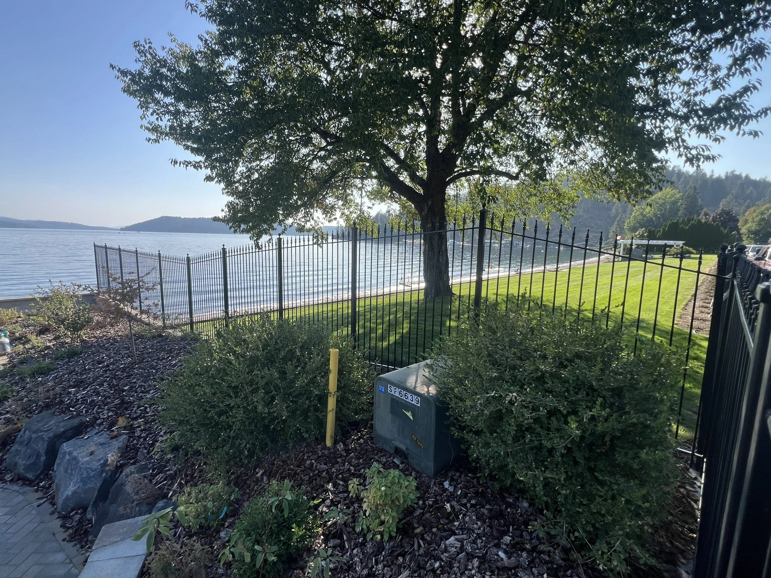 A lakeside scene with a large tree in the foreground, surrounded by bushes and rocks, a black metal fence, and a well-maintained grassy area. The lake and distant hills are visible under a clear blue sky.