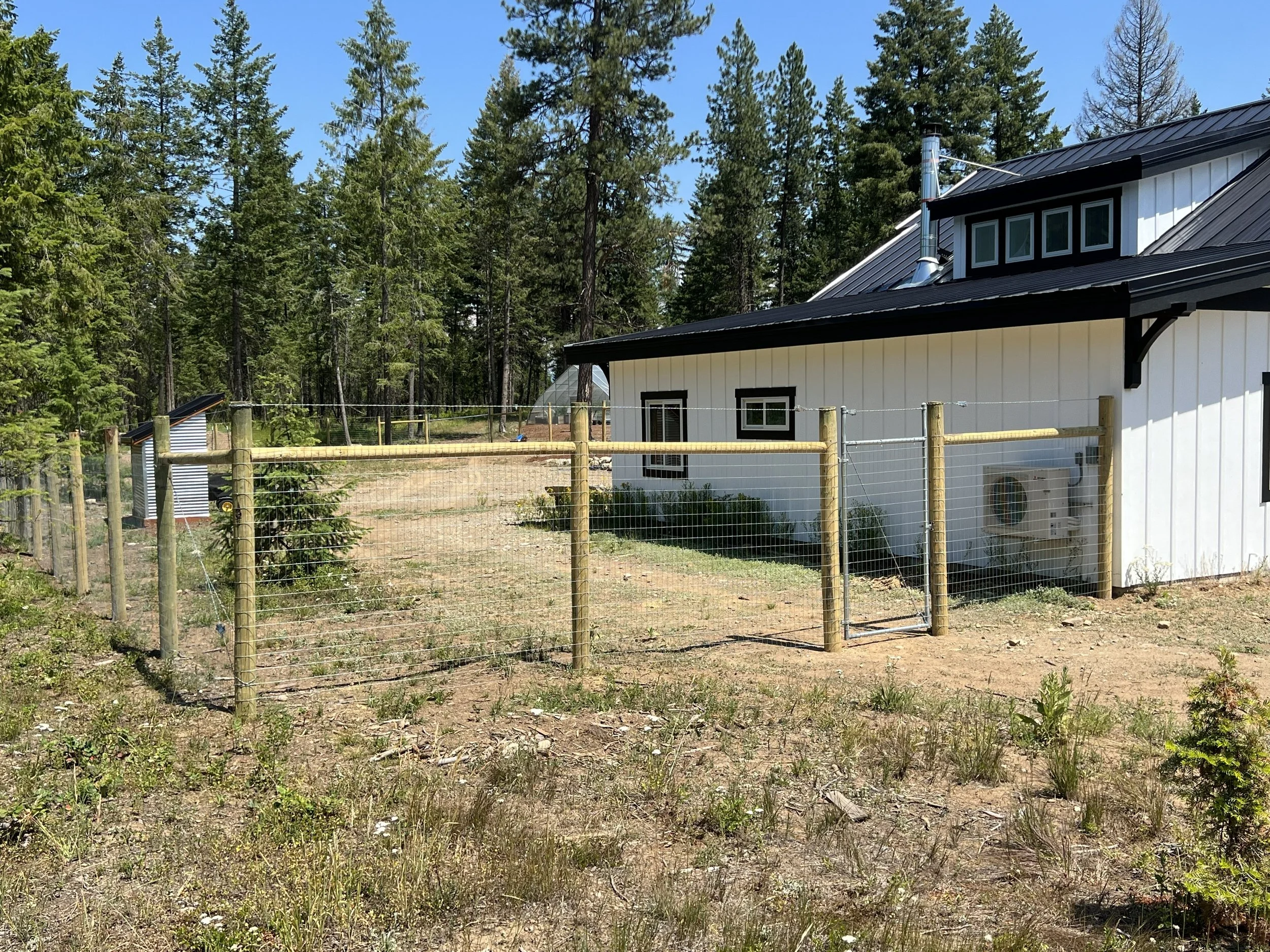 Residential house with a metal roof, white siding, and small windows, surrounded by a wire and wooden fence, with a dirt yard and a wooded area with tall trees in the background.