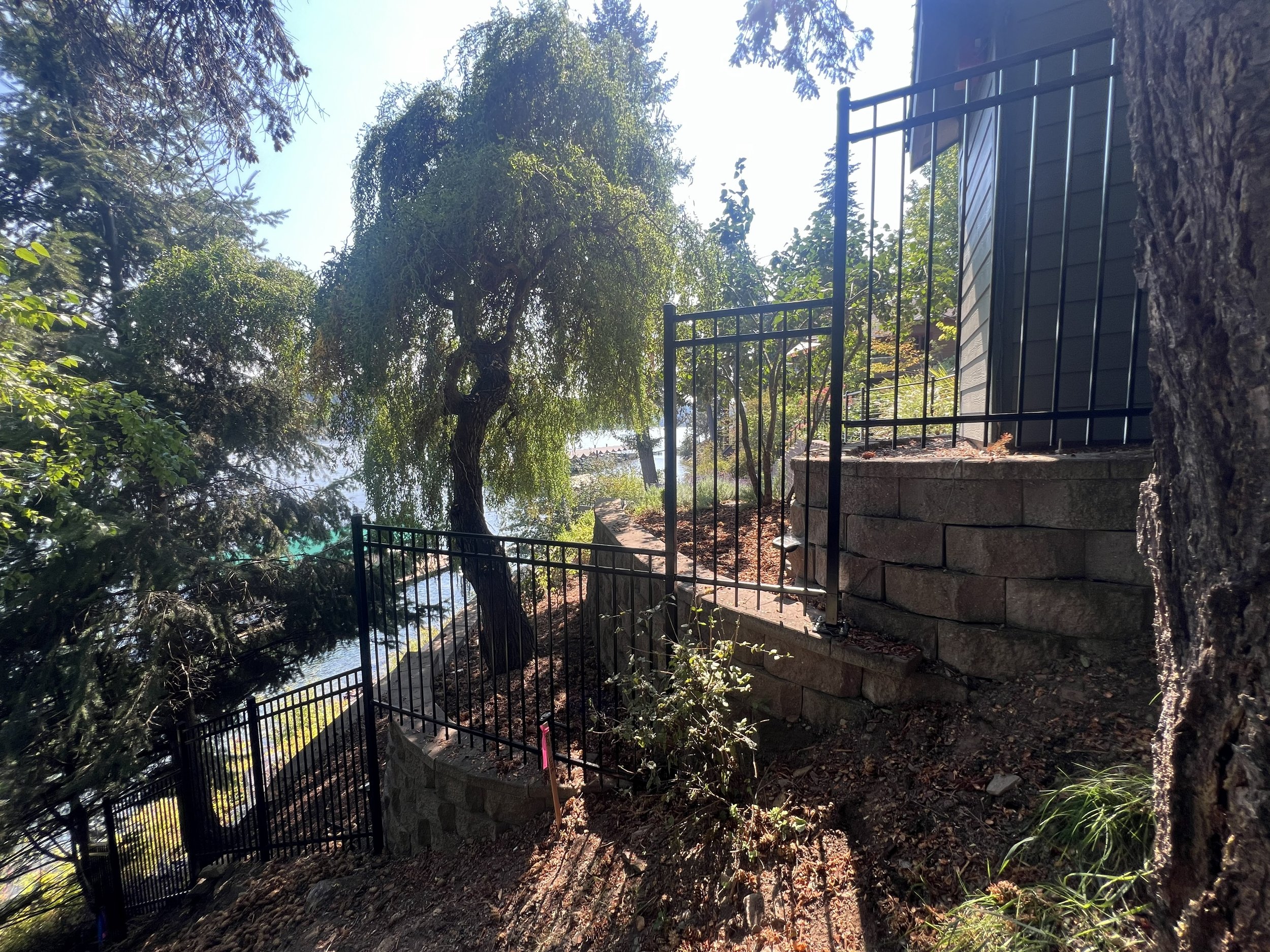 A steep backyard with stone retaining walls and black metal fencing, overlooking a body of water and surrounded by trees.