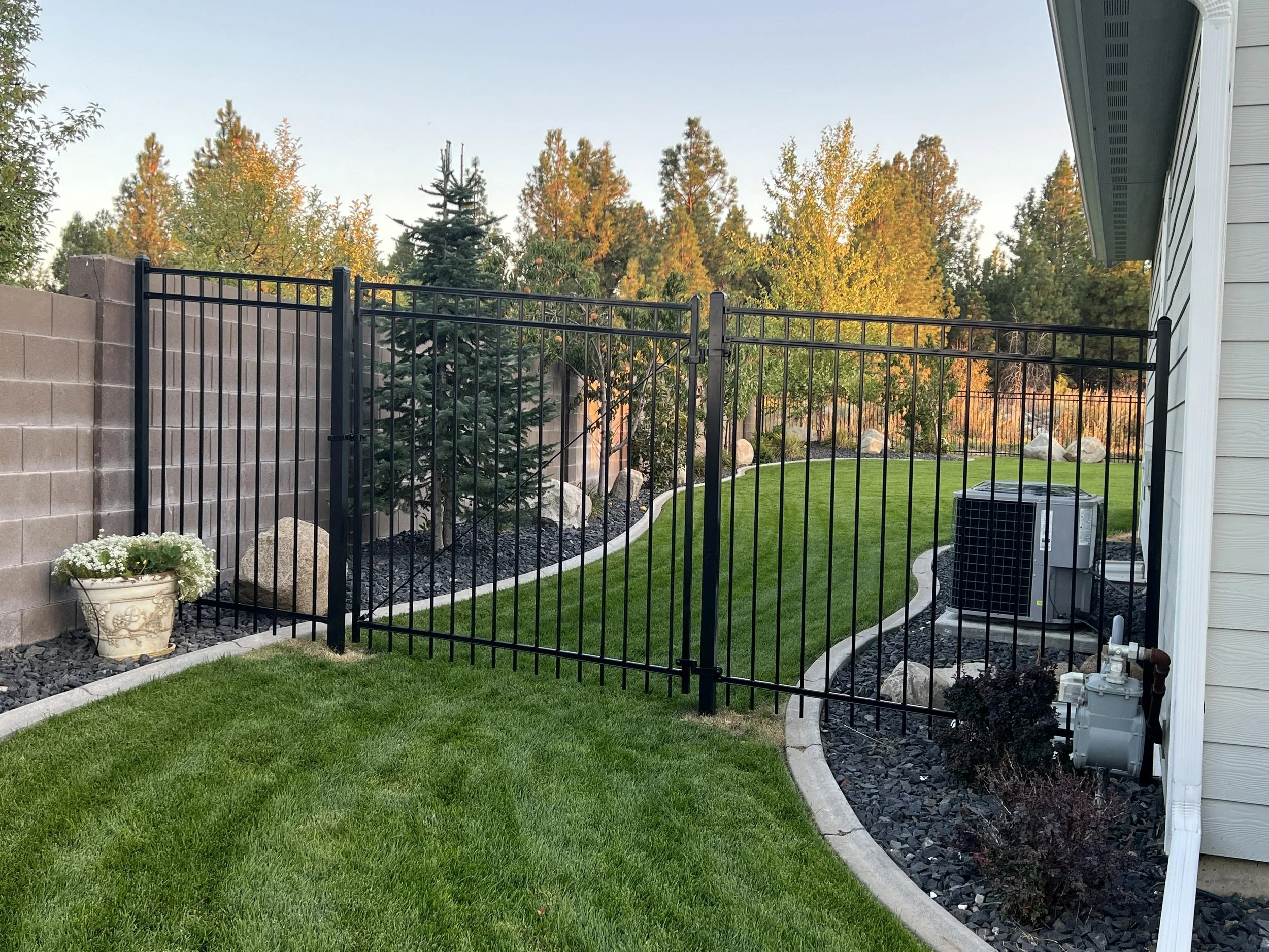 A backyard with a black metal fence, green grass, a gray stone wall, potted flowers, and outdoor air conditioning unit beside a beige house with white siding, surrounded by trees with autumn foliage.
