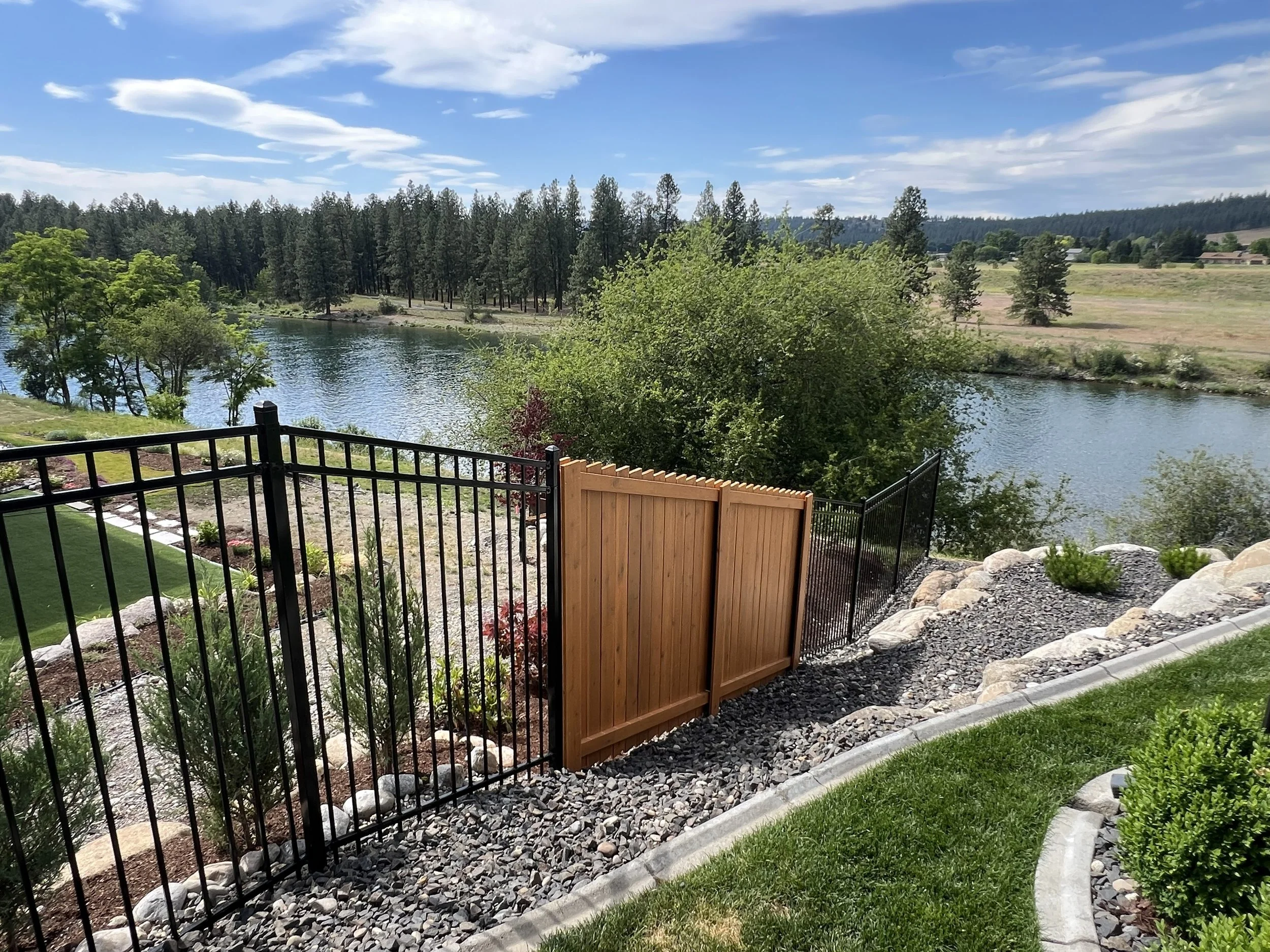 View of a lake and trees from a backyard with a black metal fence and a wooden gate, alongside rocky landscaping and some green grass