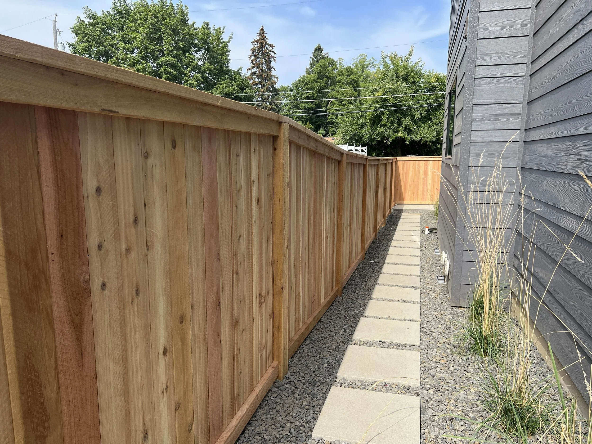 A backyard pathway with concrete stepping stones alongside a newly built wooden fence on the left and a gray house with siding on the right. There are some dry grasses growing near the house, and trees and power lines are visible in the background.