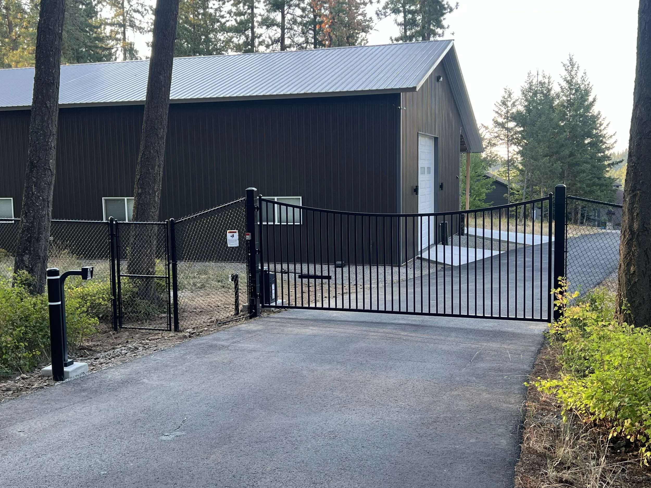 A black metal driveway gate in front of a black building with a white garage door, surrounded by trees and greenery.