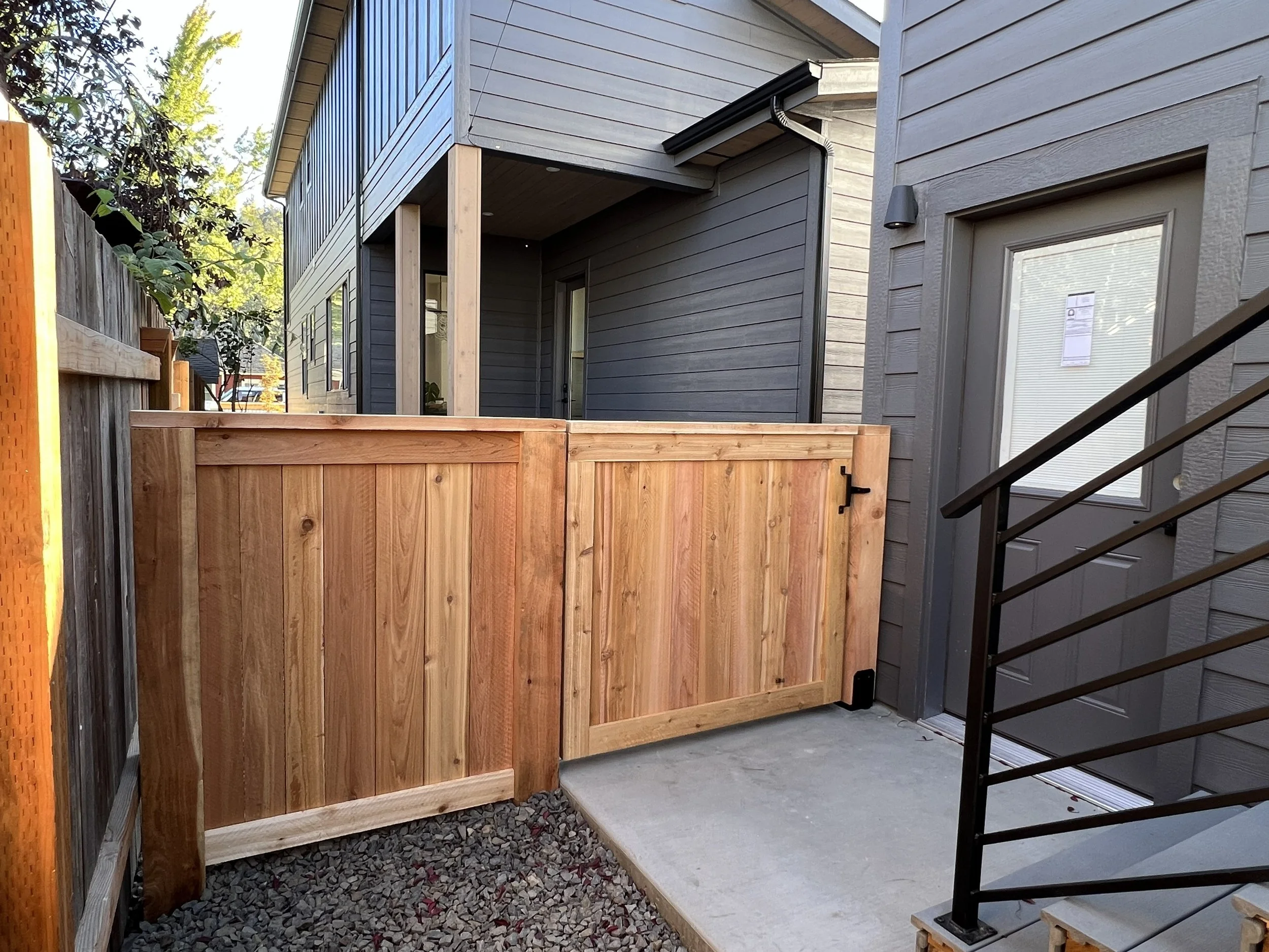 Backyard with wooden gate, concrete patio, black metal railing, and gray house with window and door.