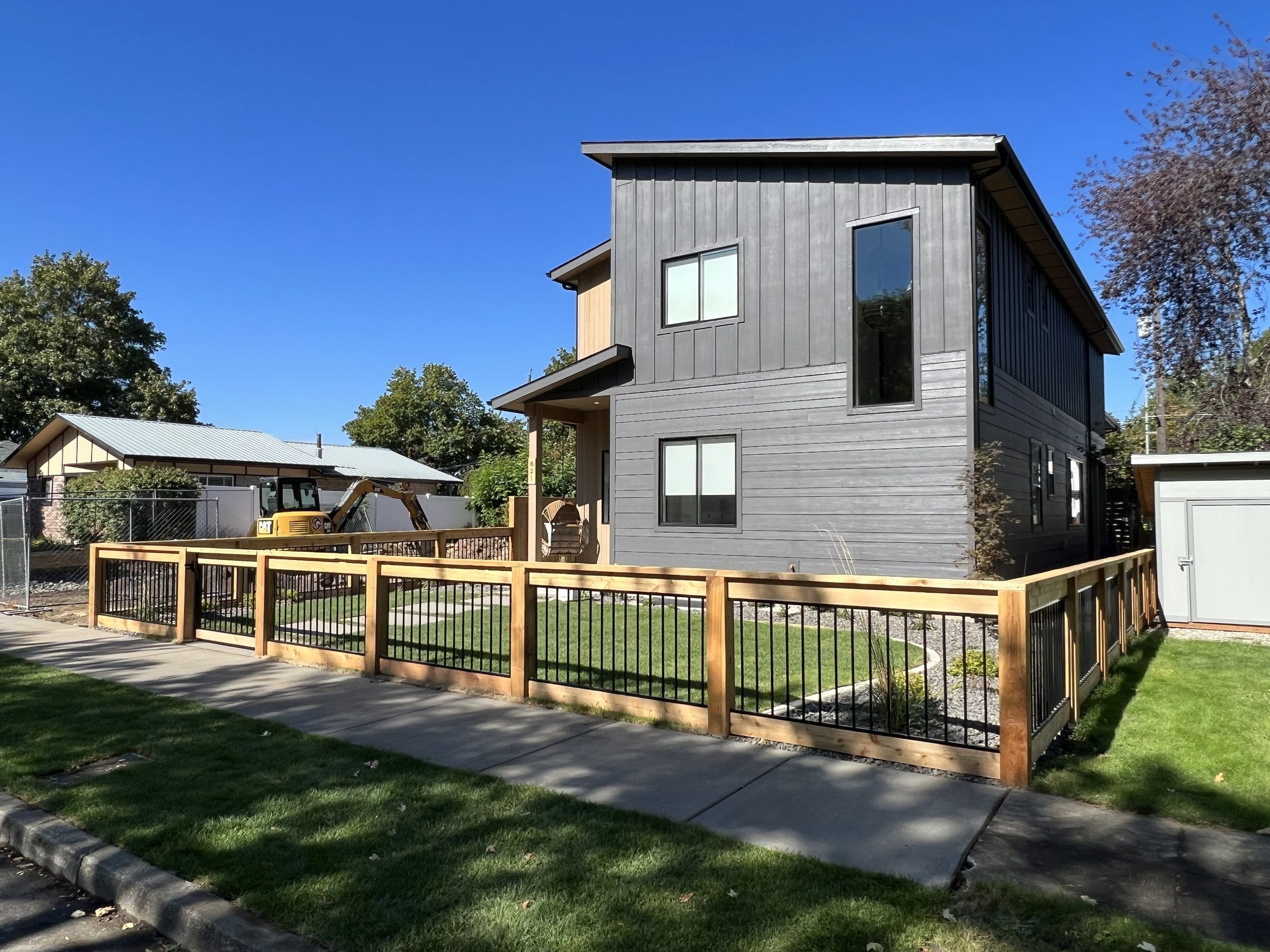 Newly constructed two-story house with dark gray siding, large windows, and a wooden fenced yard, under a clear blue sky.