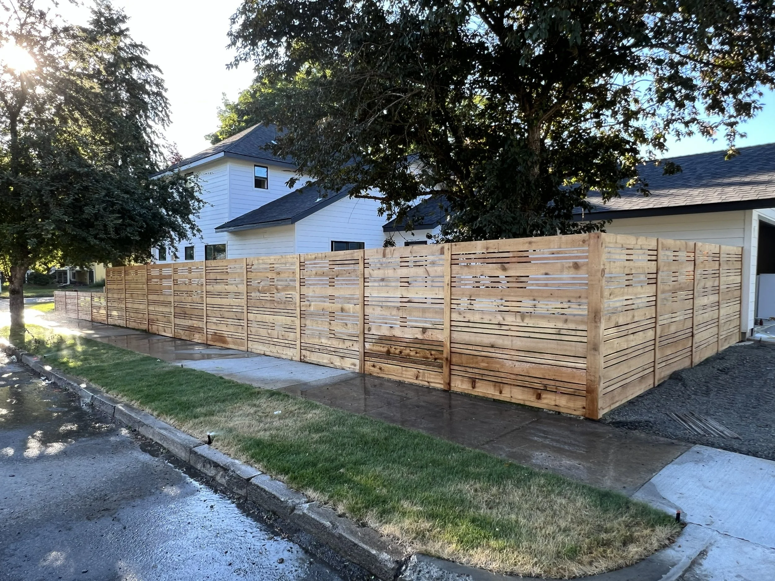 New wooden privacy fence on a residential sidewalk, with wet pavement and grass in the foreground, and trees and houses in the background.