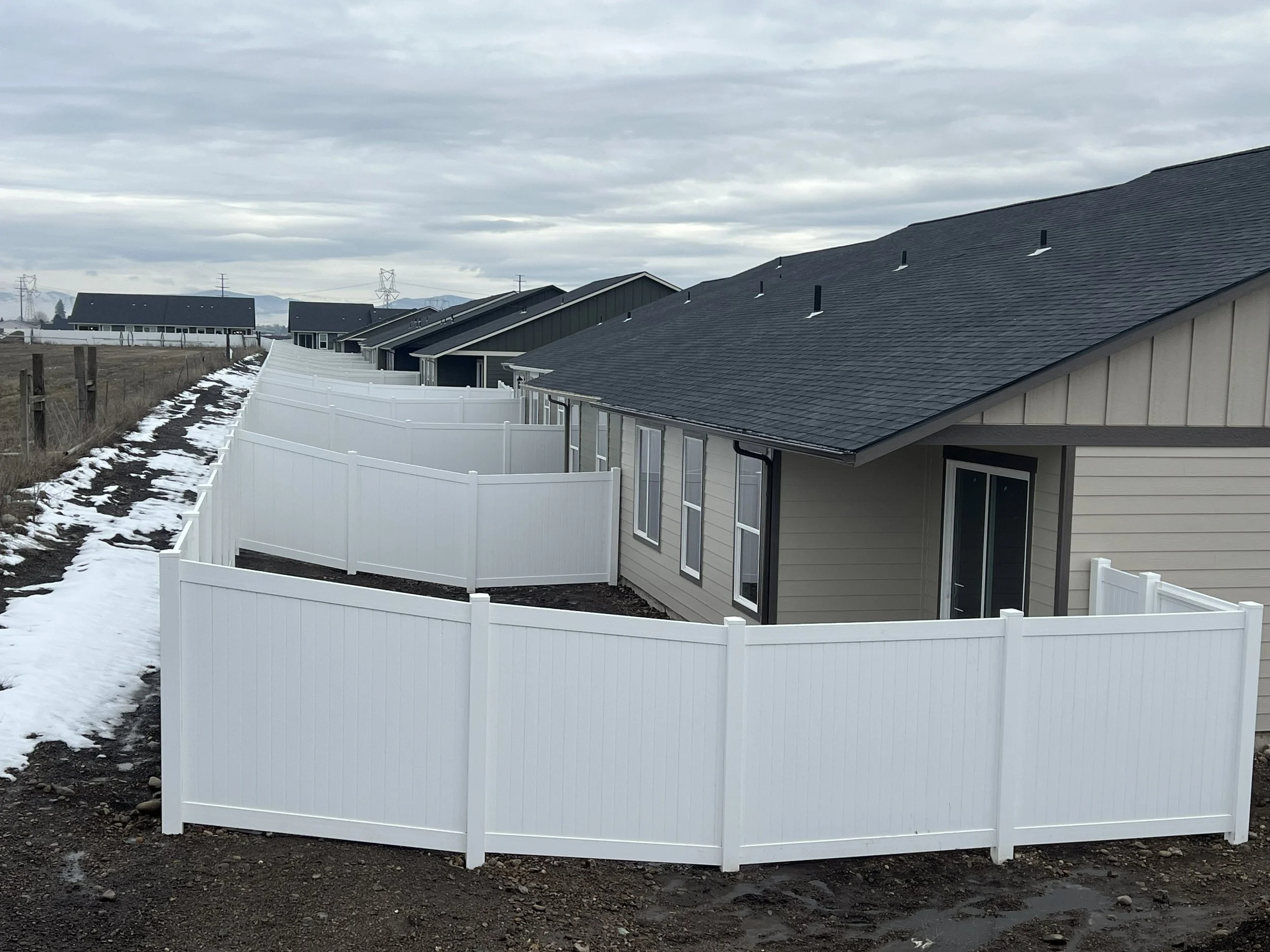 Residential houses with black roofs, beige and blue siding, white fences, and snow on the ground under a cloudy sky.
