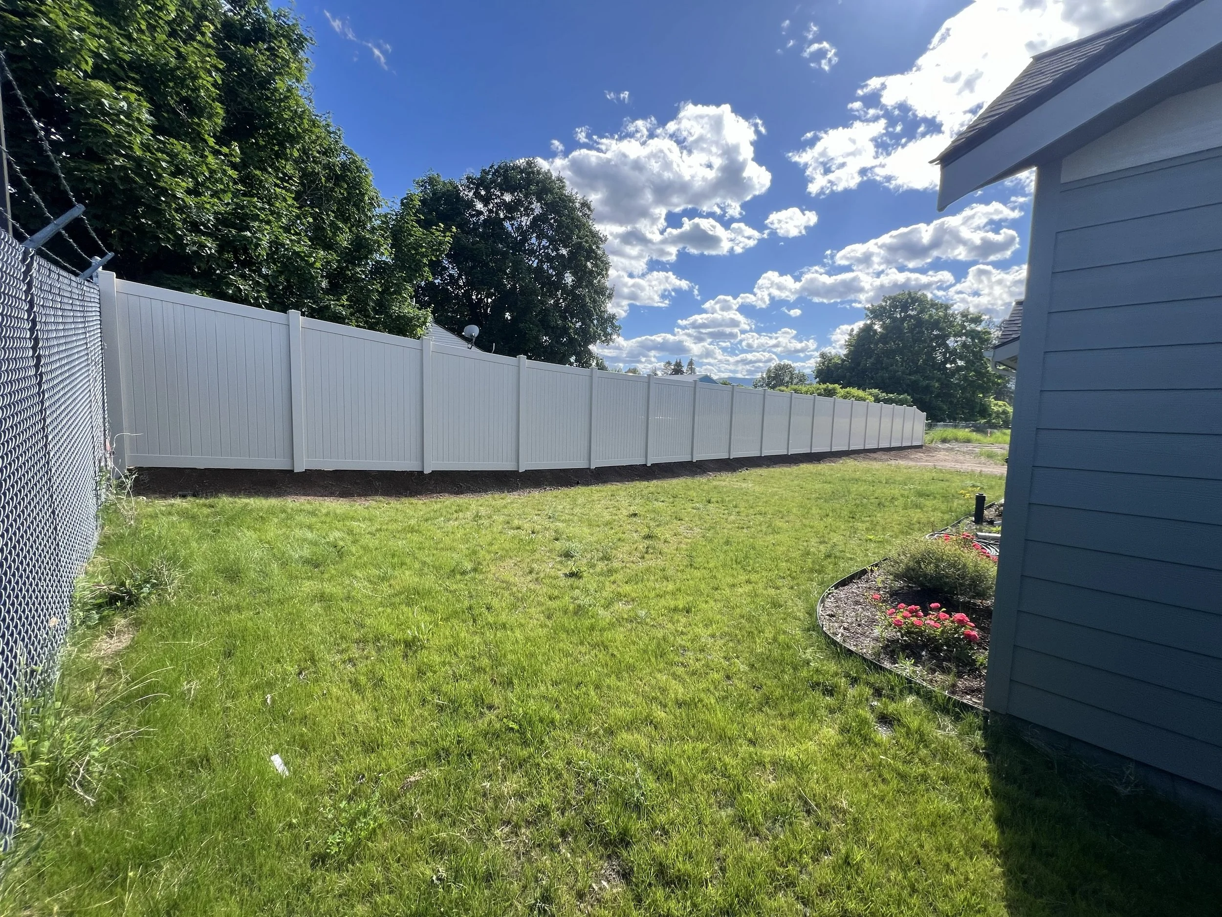 A backyard with a white vinyl fence, green grass, flower bed with pink flowers, blue house siding, and a partly cloudy sky.
