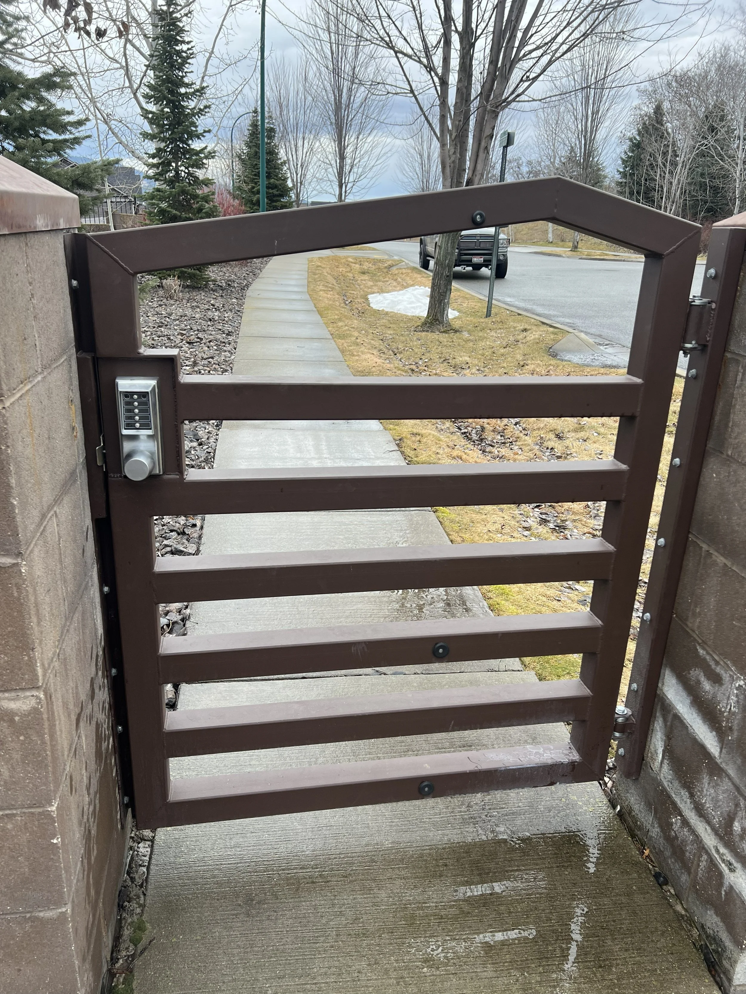 A brown metal security gate with keypad lock on a sidewalk between brick walls, overlooking a residential street with trees, parked cars, and a patch of snow.