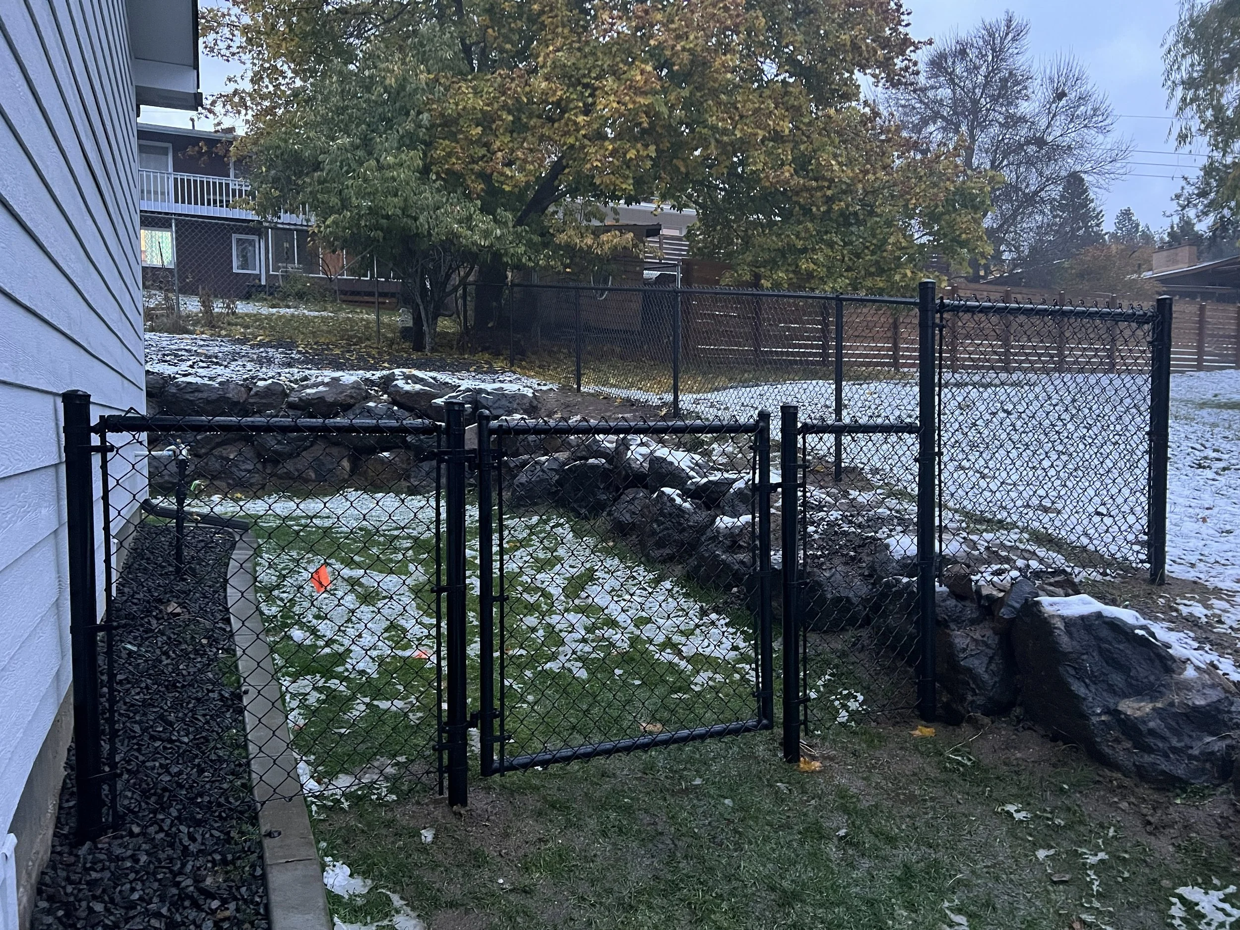 Residential backyard with black metal fence gate, stone retaining wall, patches of snow, green grass, and trees in the background during winter.