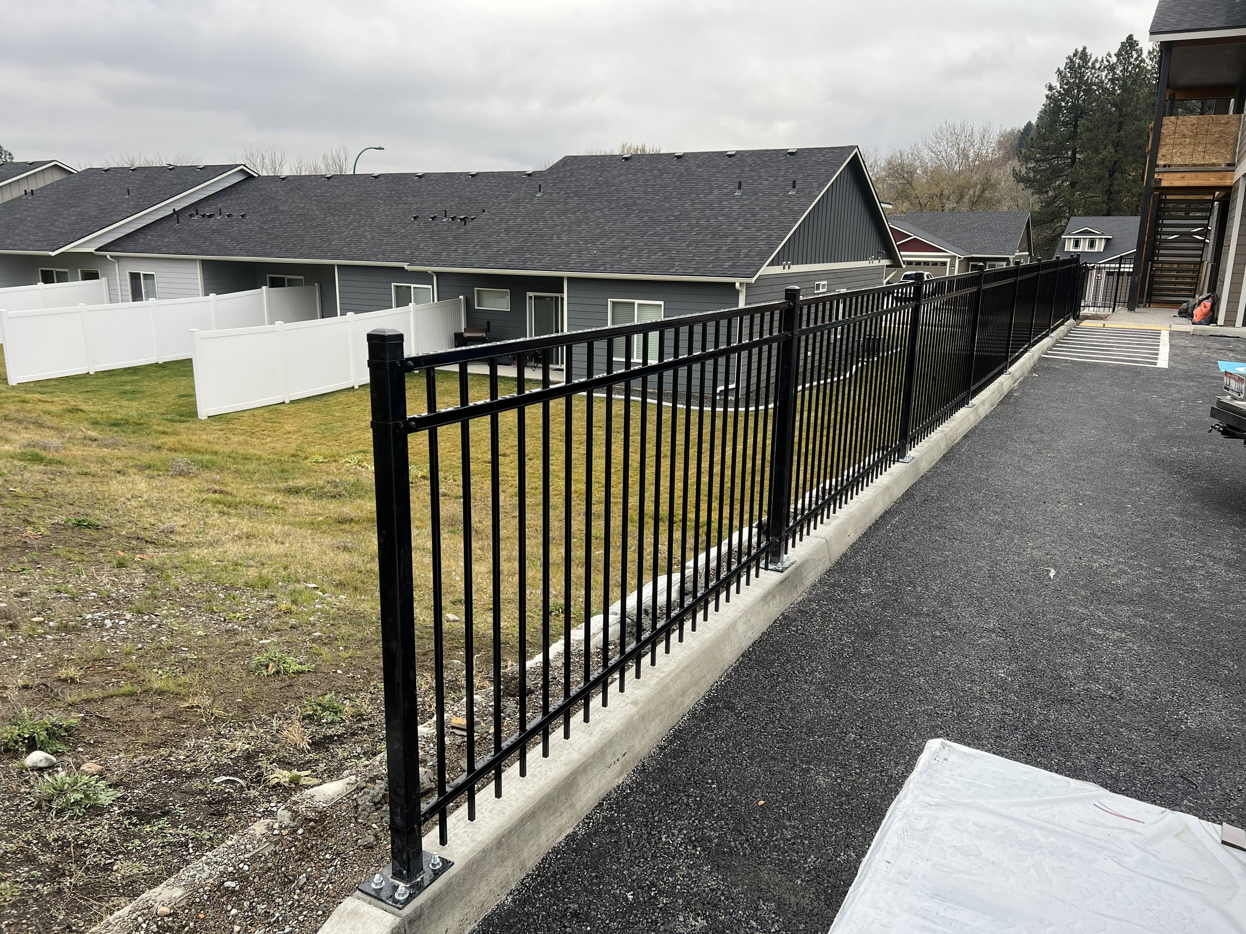 Black metal fence along a paved area next to a grassy yard with houses in the background under cloudy sky.